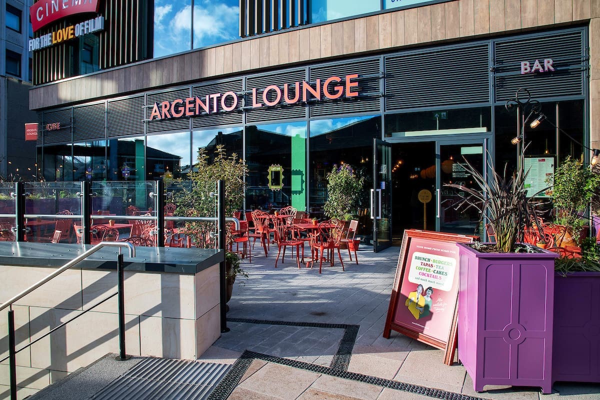 Outdoor view of Argento Lounge café with red chairs and tables on the patio, glass fence, plants, and a purple stand with a sign beside the entrance; large windows of Argento reflect blue sky and clouds.