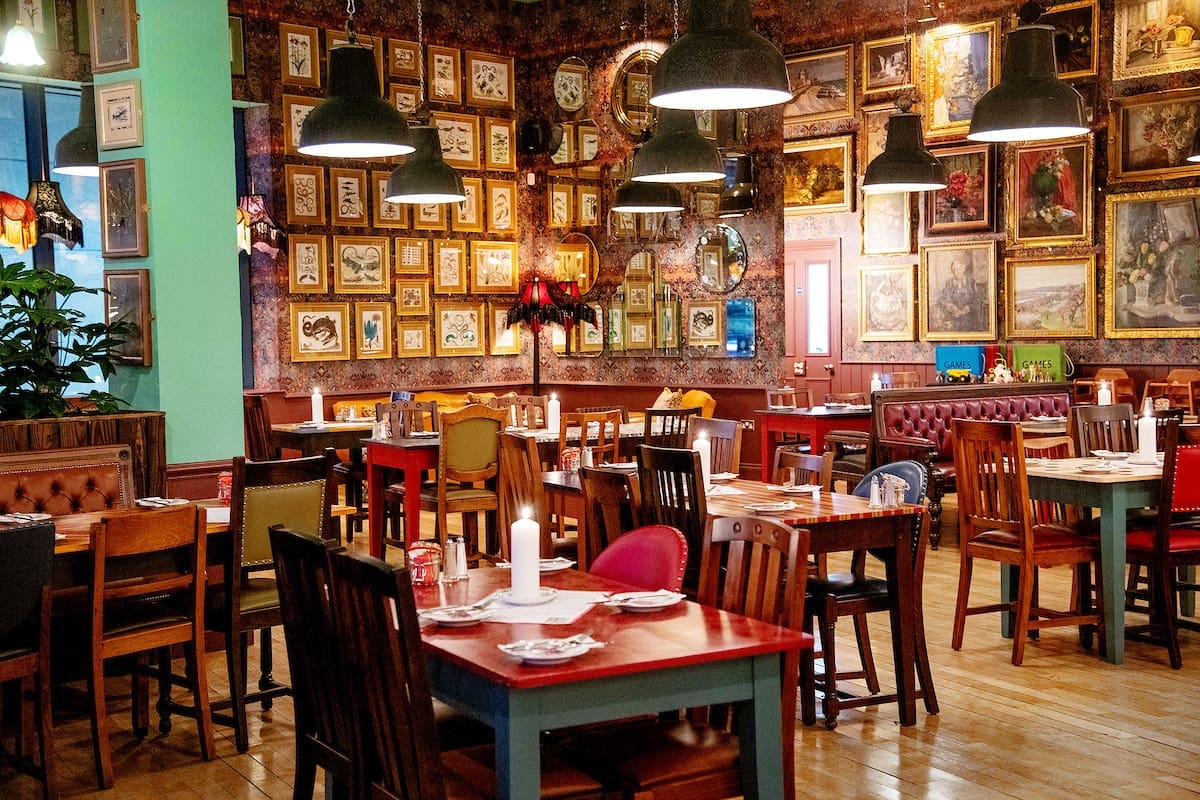 Eclectic Argento restaurant interior with wooden tables and chairs, vintage decor, and numerous framed pictures covering the walls. Warm lighting hangs from the ceiling, creating a cozy and inviting atmosphere.