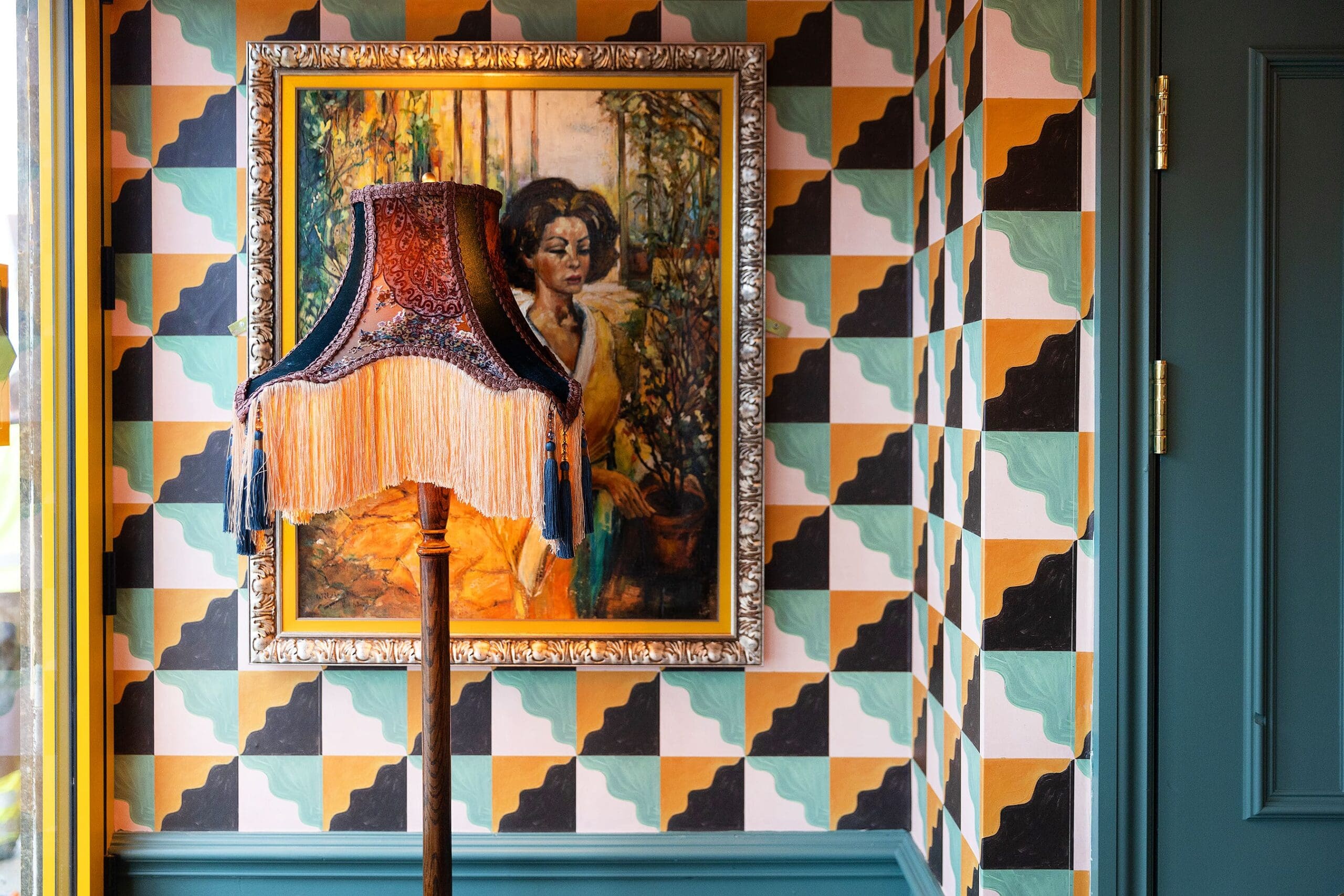 A vintage fringed lamp stands in front of a framed painting of a woman, set against a vibrant manzano geometric-patterned wall in teal, black, peach, and white tones.