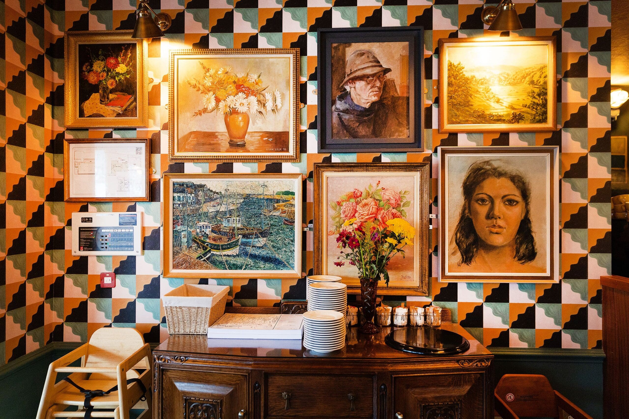 A wooden sideboard with flowers, dishes, and condiments stands against a patterned wall decorated with framed paintings of portraits, landscapes, manzano blossoms, and flowers in a cozy, warmly lit room.