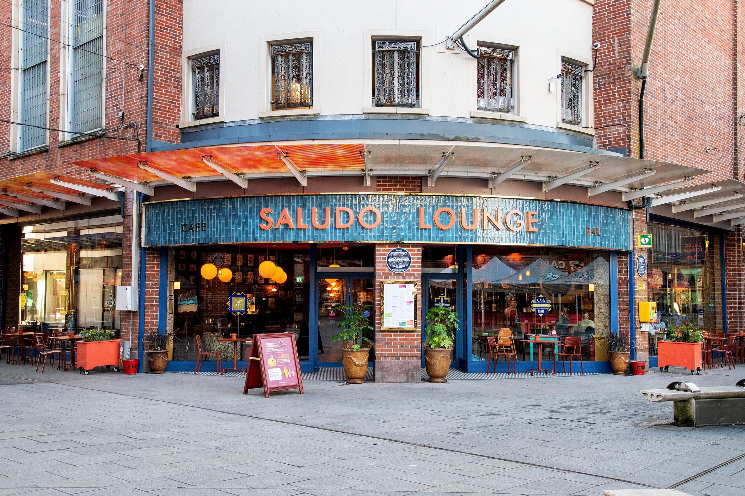 The exterior of Saludo Lounge café features outdoor seating, colorful chairs and tables, potted plants, and large windows on a city street corner. The building has a curved brick façade with a welcoming saludo atmosphere.