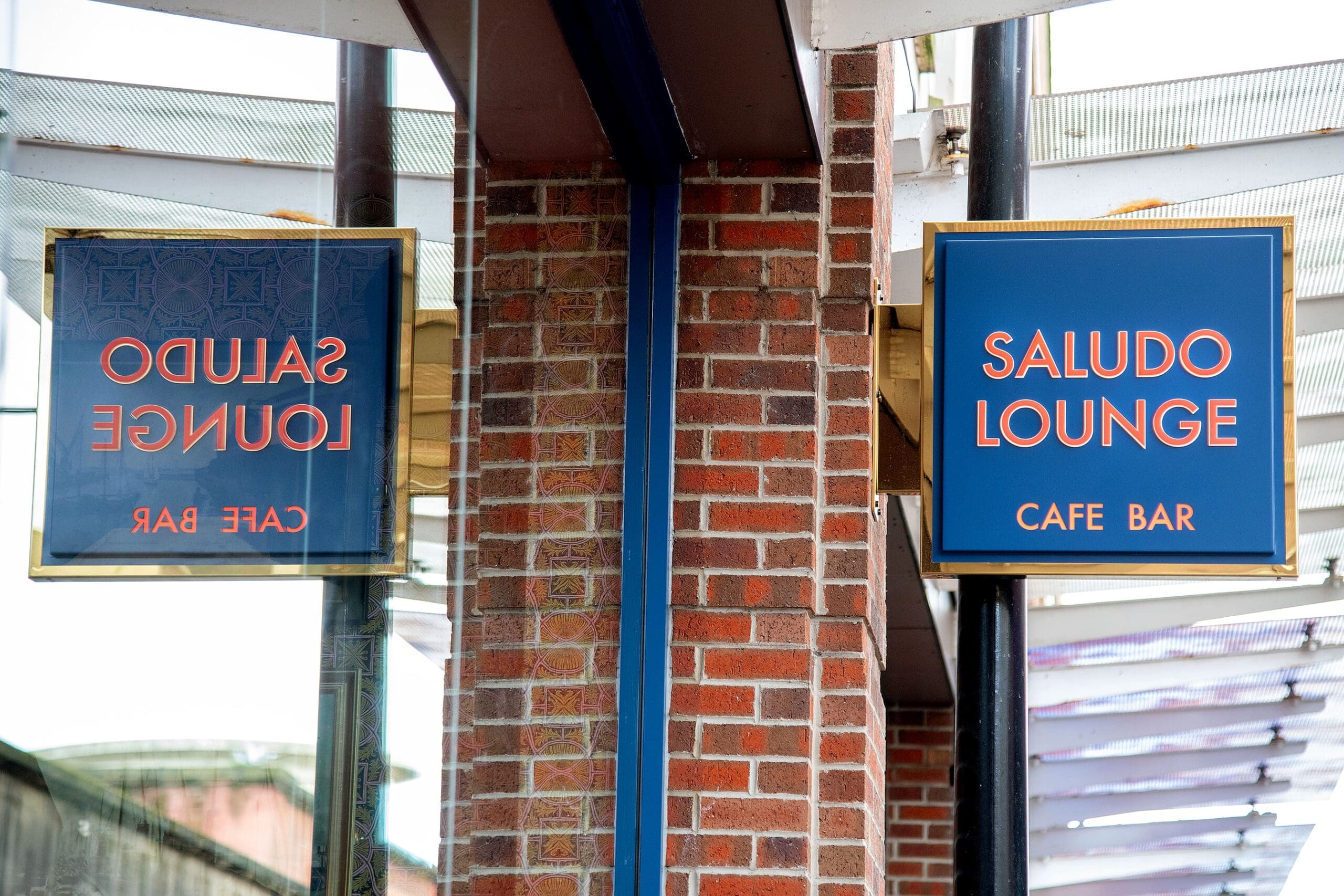 A blue saludo sign with gold trim reads "SALUDO LOUNGE CAFE BAR" next to a brick wall. The sign is reflected in glass, displaying the reversed text.