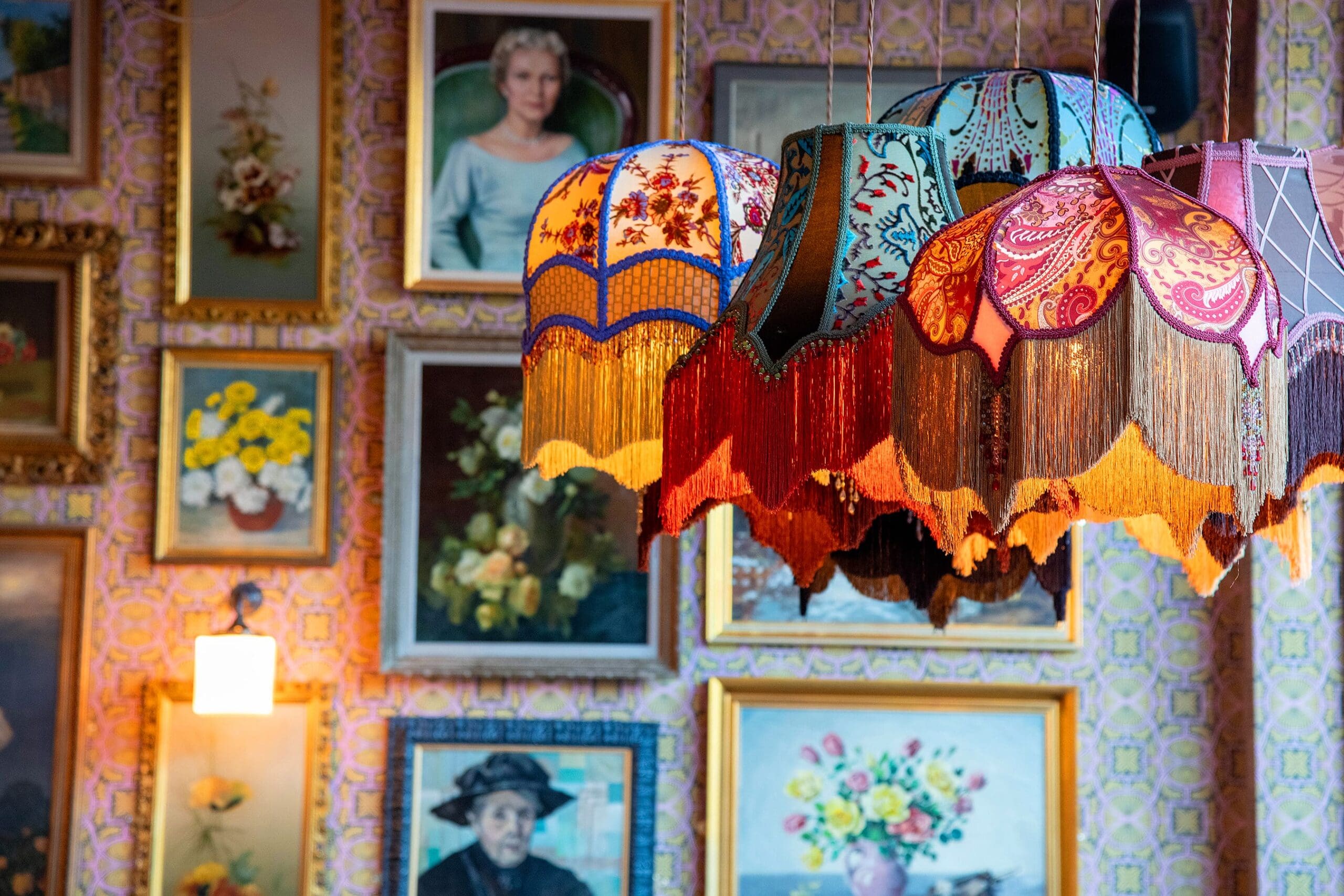 Colorful, vintage lampshades with tassels hang from the ceiling in front of a wall covered in assorted framed paintings and portraits, giving the space a cozy, eclectic atmosphere that feels like a warm saludo to creativity.