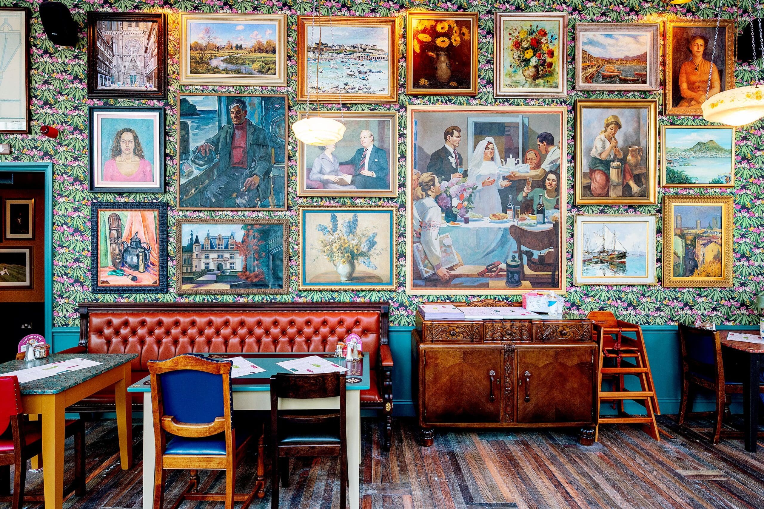 A vibrant restaurant interior with floral wallpaper, wooden floors, and a red tufted bench welcomes guests with a warm saludo. The wall is filled with framed paintings of landscapes, portraits, and still lifes above tables and chairs.