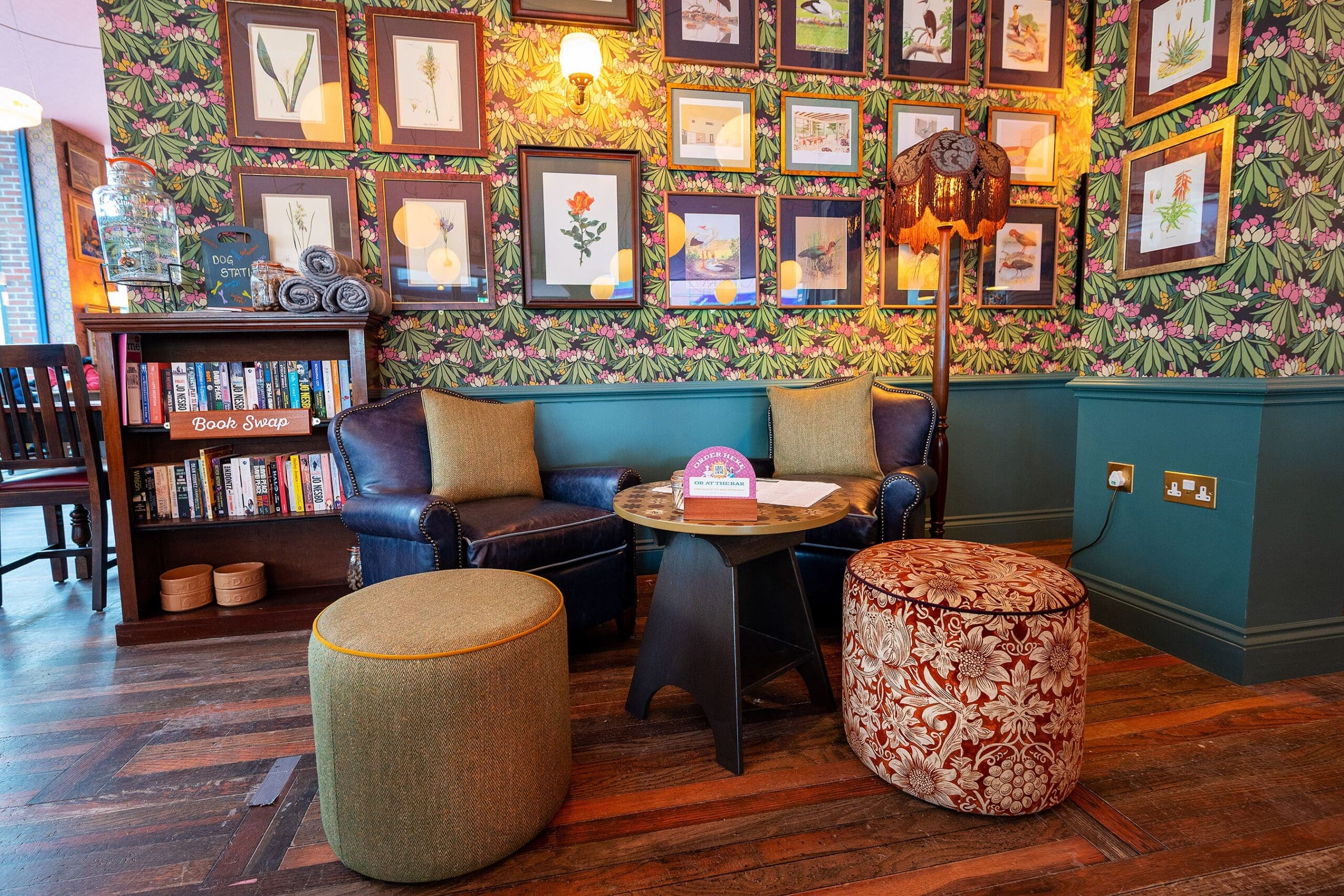 A cozy reading nook with a dark leather sofa, two round ottomans, a small table, lamp, bookshelf, and a wall covered in framed botanical prints delivers a warm saludo against floral wallpaper and wooden flooring.