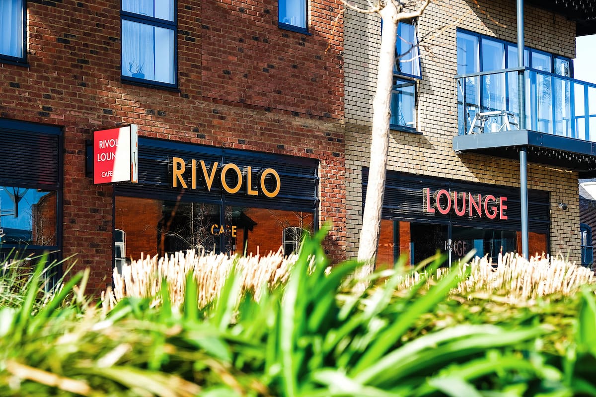 A café named "Rivolo Lounge" with its bold red and yellow signage brightens a brick building, partially hidden behind tall green grass on a sunny day. The inviting spot reflects Rivolo’s signature charm.