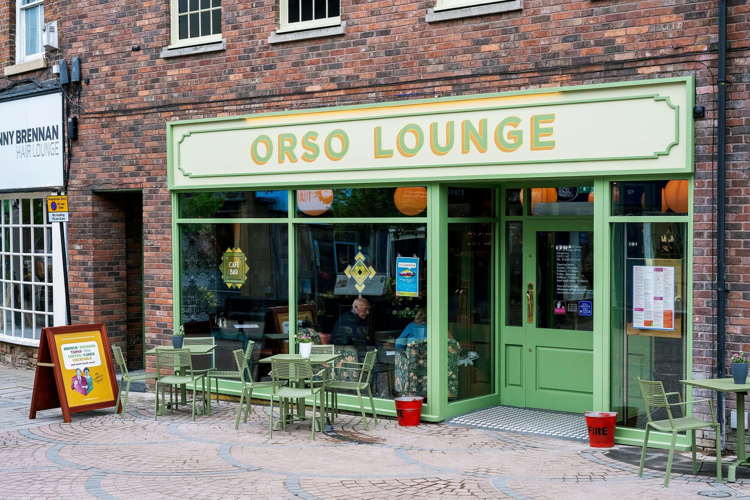 Street view of Orso Lounge, a cozy cafe with green-framed windows and doors. Green outdoor tables and chairs line the stone pavement, while a red sandwich board welcomes you to Orso near the entrance.