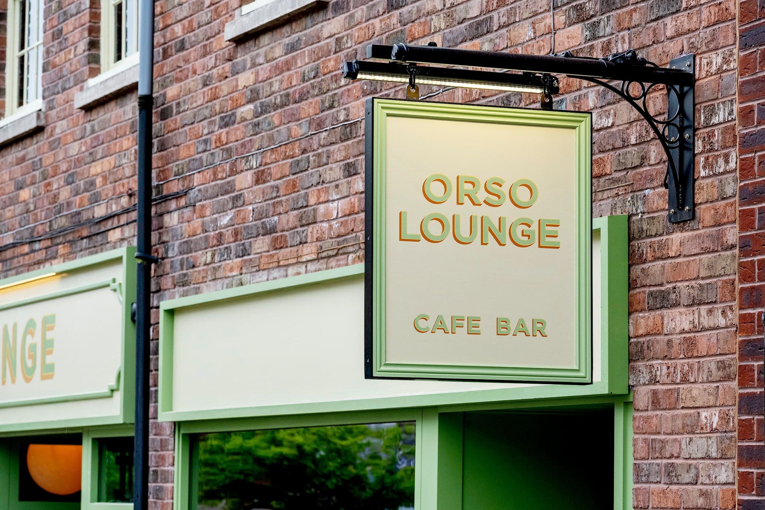 A green and gold sign reading "Orso Lounge Cafe Bar" hangs outside a brick building above a window. The facade features light green trim, adding charm to the inviting Orso exterior.
