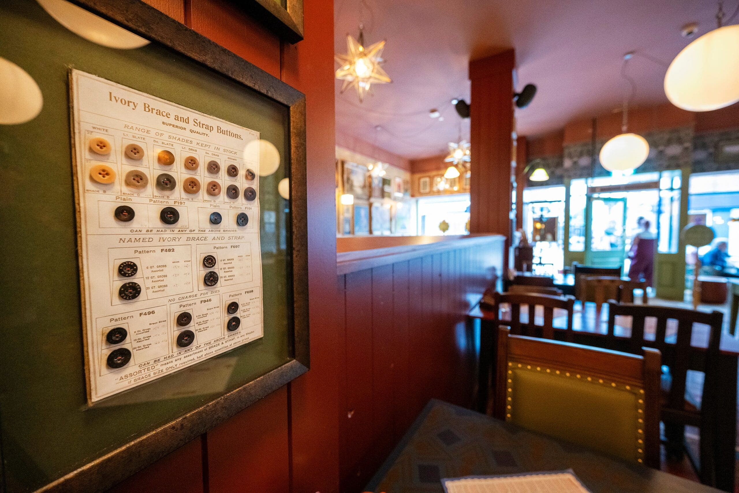 A cozy orso cafe interior with wooden tables and chairs, warm lighting, and hanging star-shaped lamps. A framed display of vintage buttons is mounted on a red wall, and a person stands near the entrance in the background.
