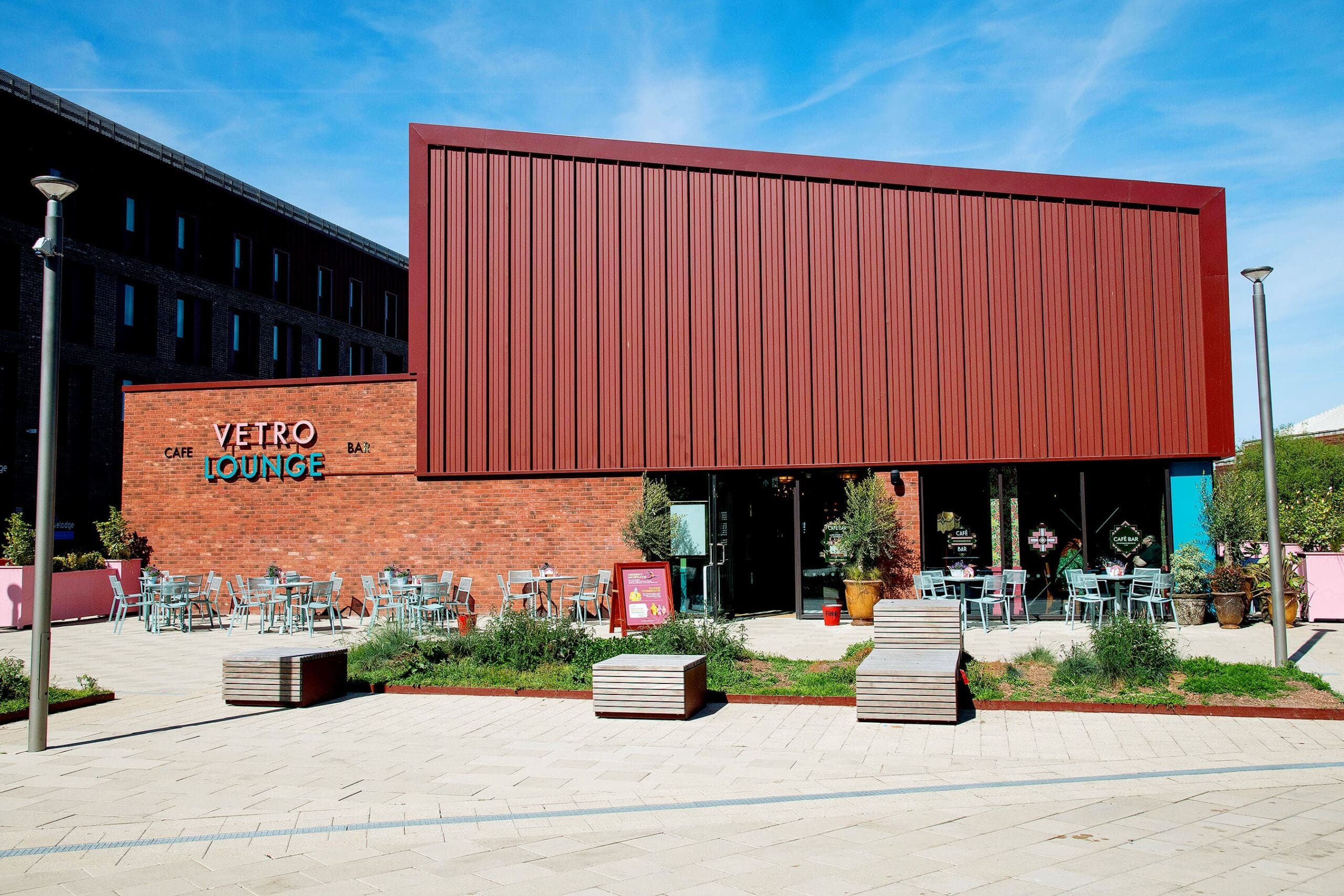 Modern café-bar with a red angular facade and large windows, labeled "Vetro Lounge." Outdoor Vetro seating with metal tables and chairs lines a paved area, surrounded by greenery under a bright blue sky.