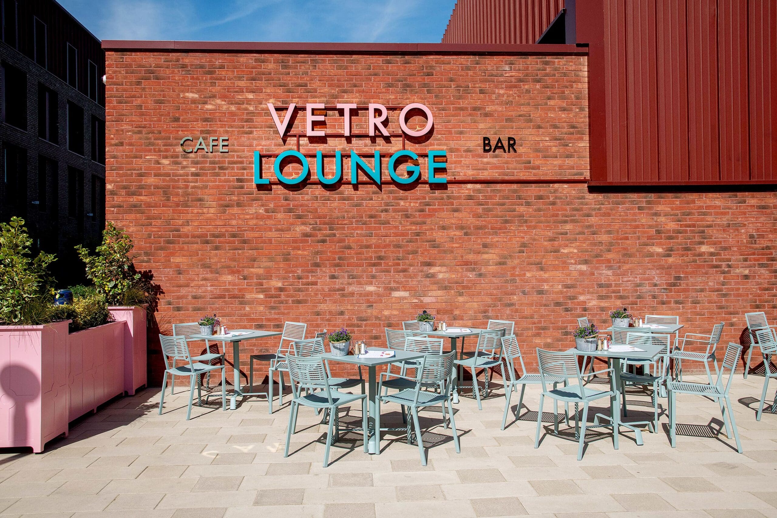 Outdoor seating area with mint green chairs and round tables in front of a red brick wall featuring the Vetro Lounge sign, along with “Cafe” and “Bar.” Pink planters filled with green plants add charm to the inviting Vetro space.