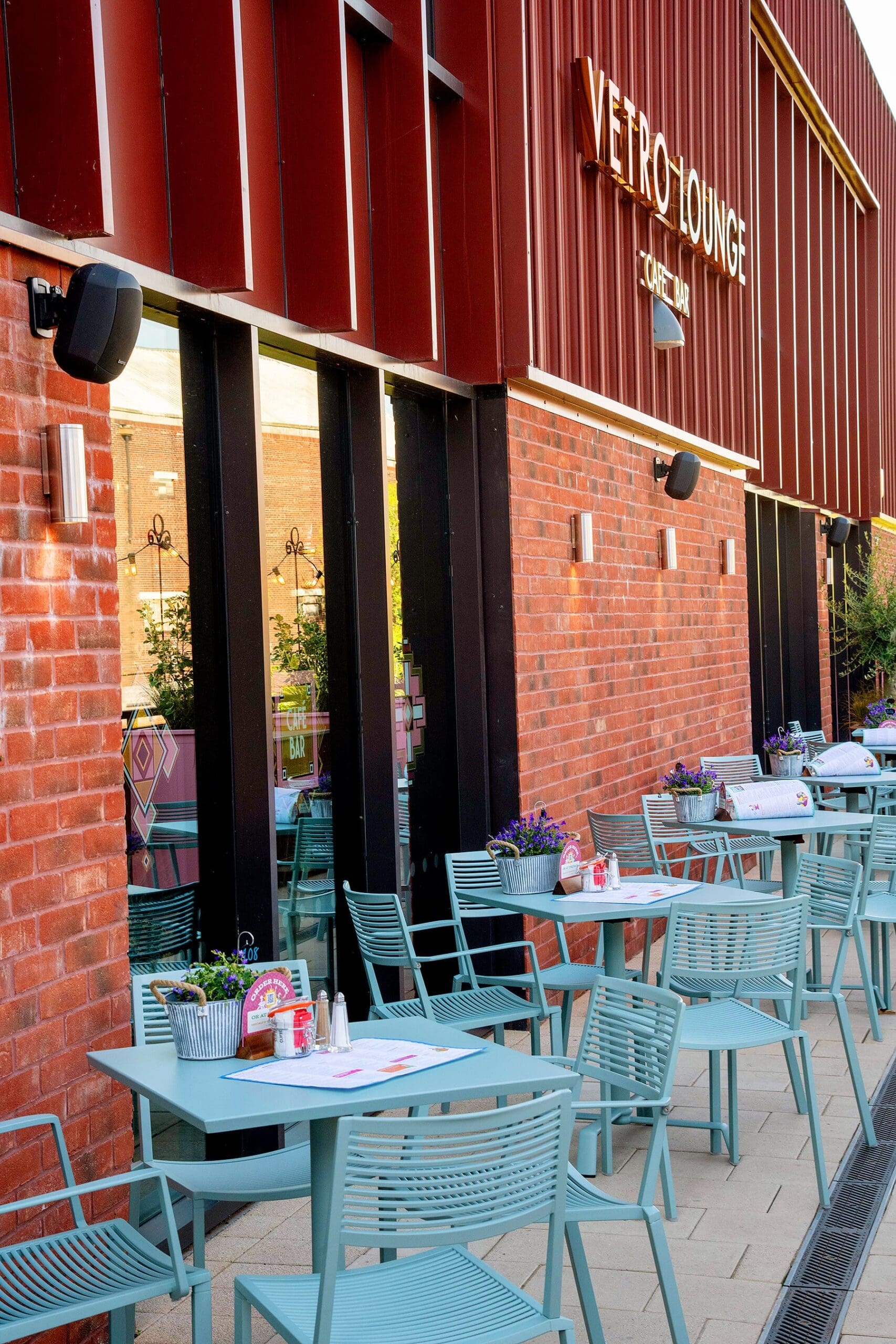 The Vetro outdoor seating area features light blue tables and chairs, potted flowers on each table, and a brick wall with large windows. The "Vetro Lounge" sign above glows in the sunlight, creating a warm and inviting atmosphere.