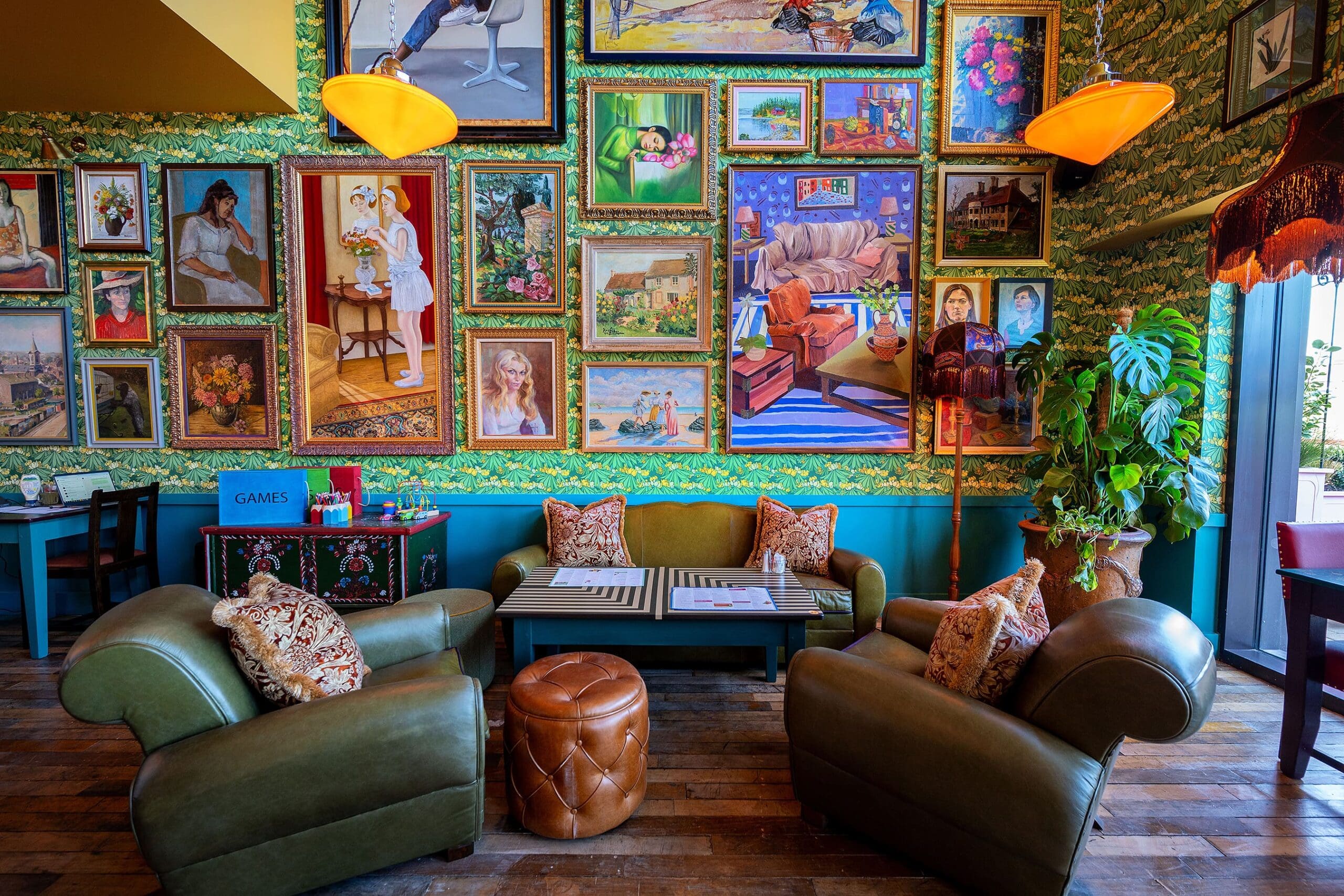 Colorful art-filled cafe with green armchairs, patterned pillows, and a Vetro coffee table. A wall of eclectic framed paintings, wooden floors, leafy wallpaper, and warm lighting create a cozy, vibrant atmosphere.