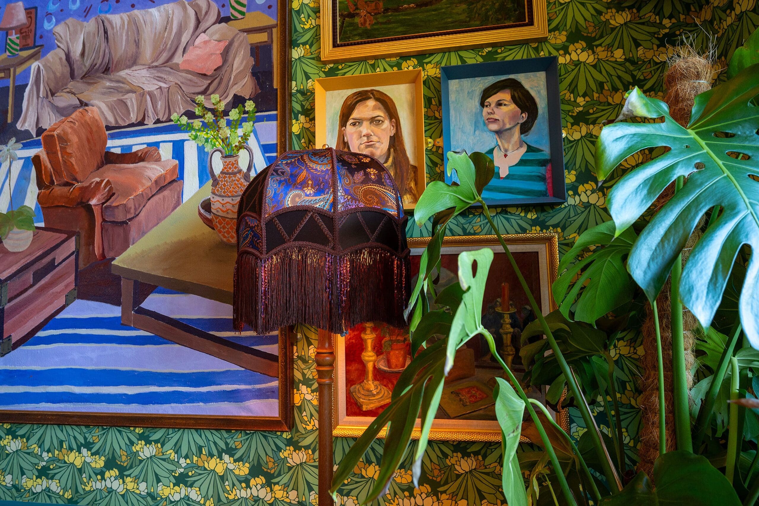 A colorful room with floral wallpaper, several framed portraits on the wall, a fringed lampshade, Vetro accents, and large green monstera leaves in the foreground.