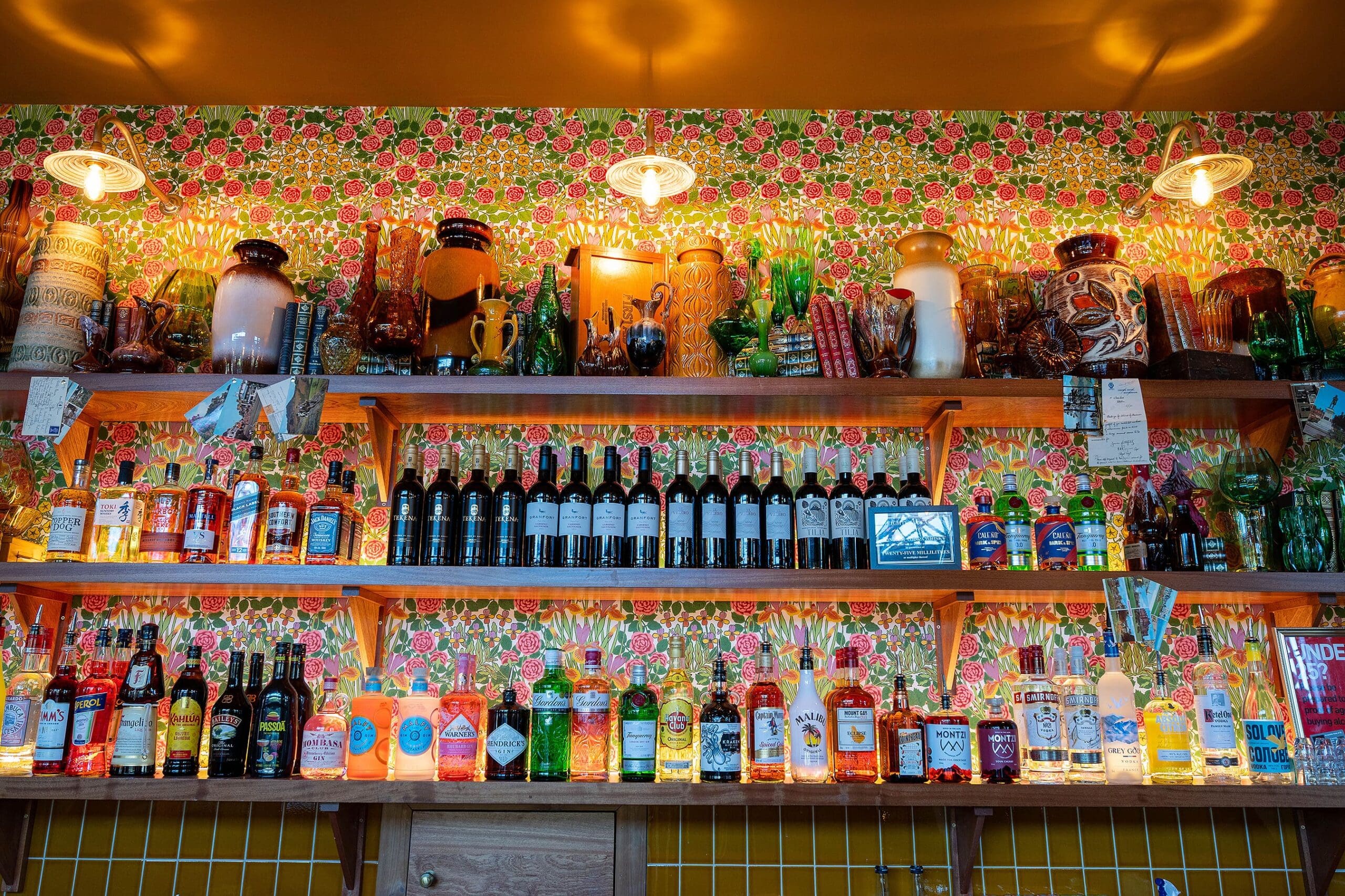 Shelves filled with assorted liquor bottles, wine, and decorative Vetro glassware, set against a colorful floral wallpaper with warm lighting from three wall-mounted lamps above.