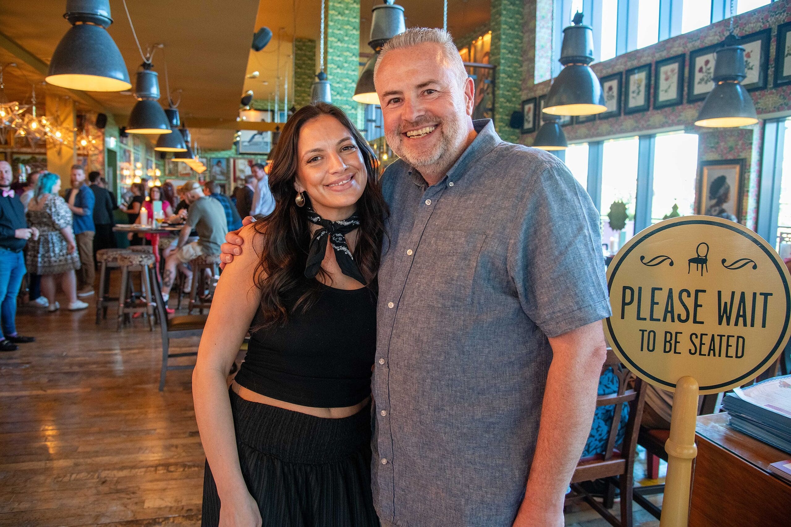 A smiling woman and man stand close together inside Vetro, a lively restaurant near a "Please Wait to Be Seated" sign, with other people and tables visible in the background.