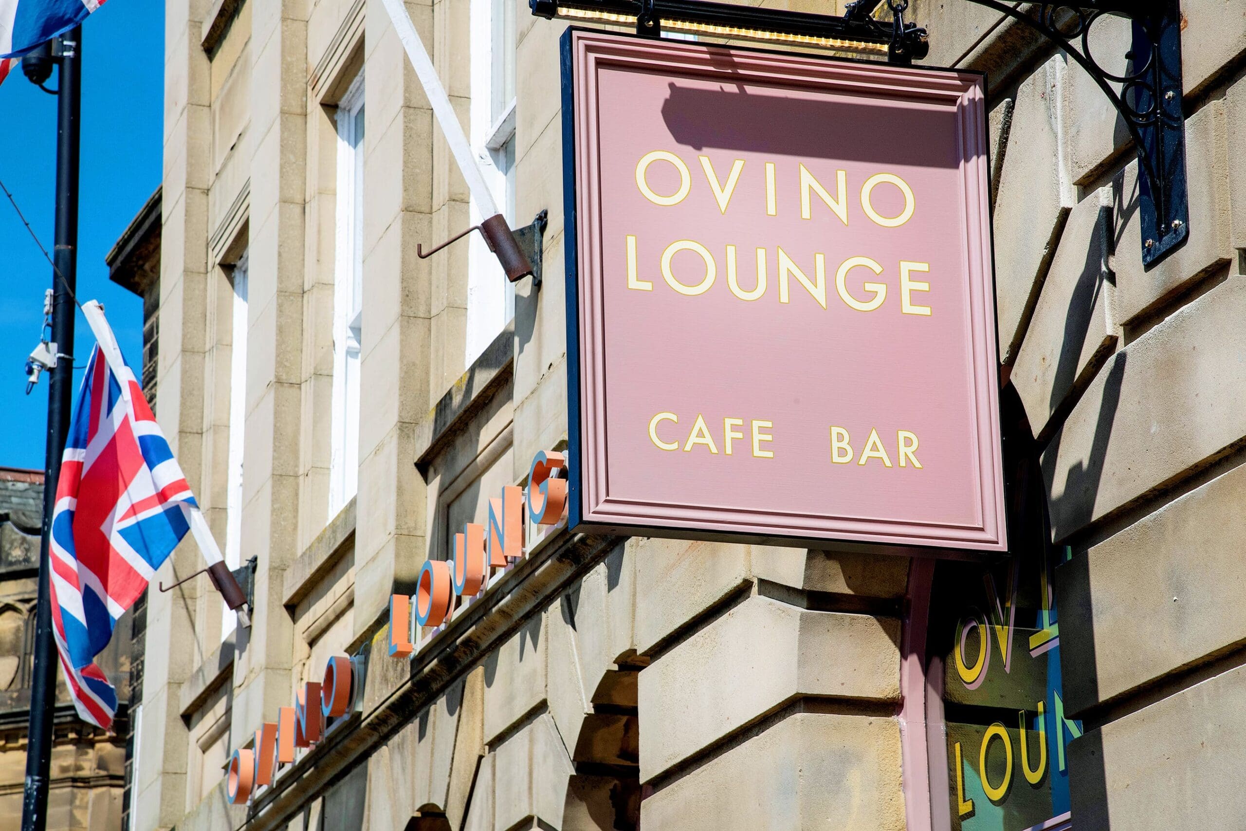 A pink Ovino Lounge Cafe Bar sign hangs outside a stone building, with the British flag to the left and bright sunlight casting shadows on the facade.