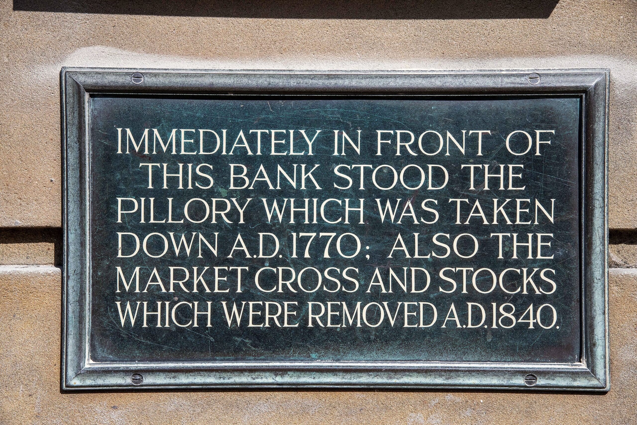 A metal plaque on a stone wall reads: "Immediately in front of this bank stood the pillory which was taken down A.D. 1770; also the market cross and stocks, once used during ovino markets, which were removed A.D. 1840.