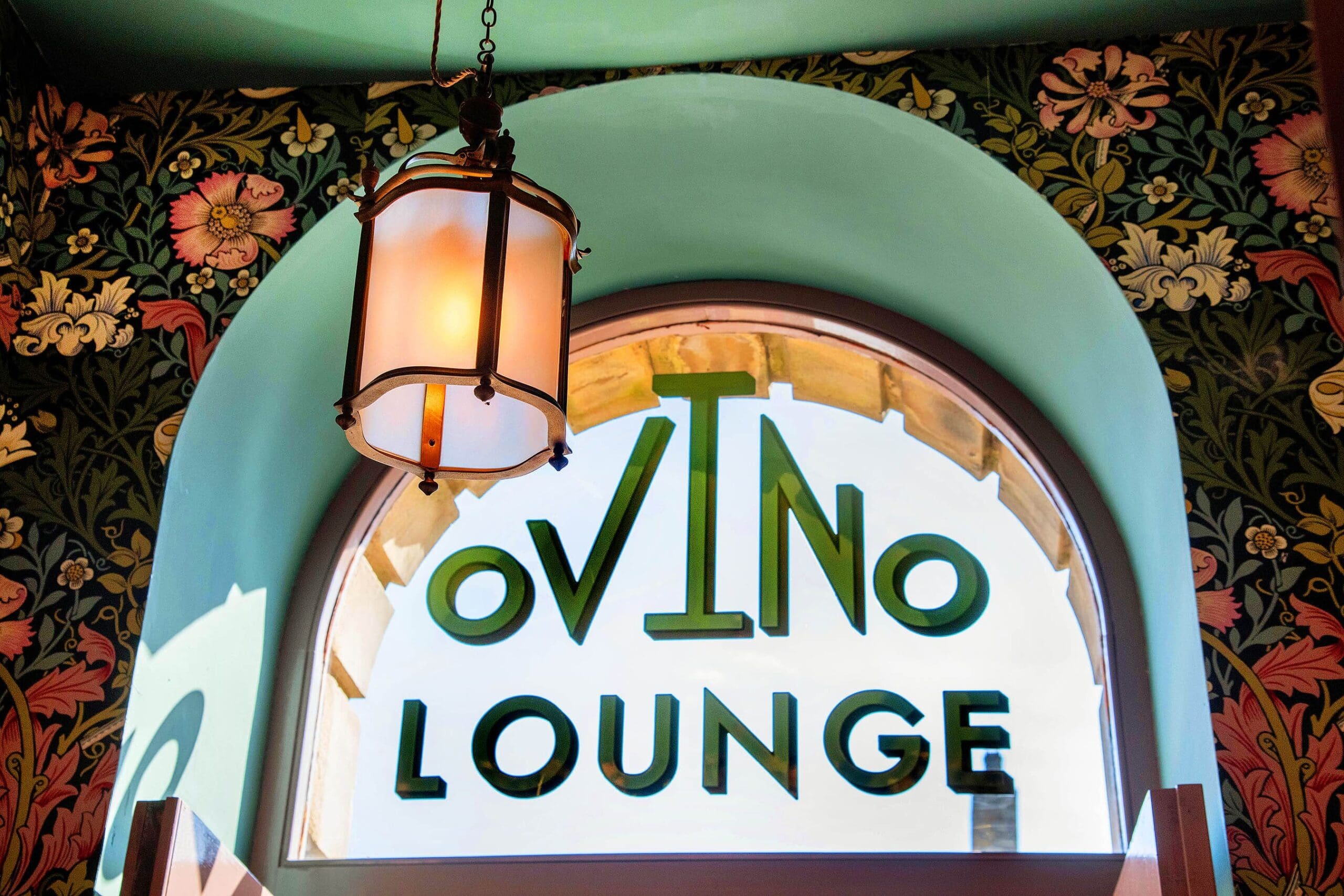 A decorative arched window with "OVINO LOUNGE" painted on it, framed by floral wallpaper and lit by a vintage hanging lantern. Sunlight streams through the glass, casting a warm glow that highlights the inviting Ovino ambiance.