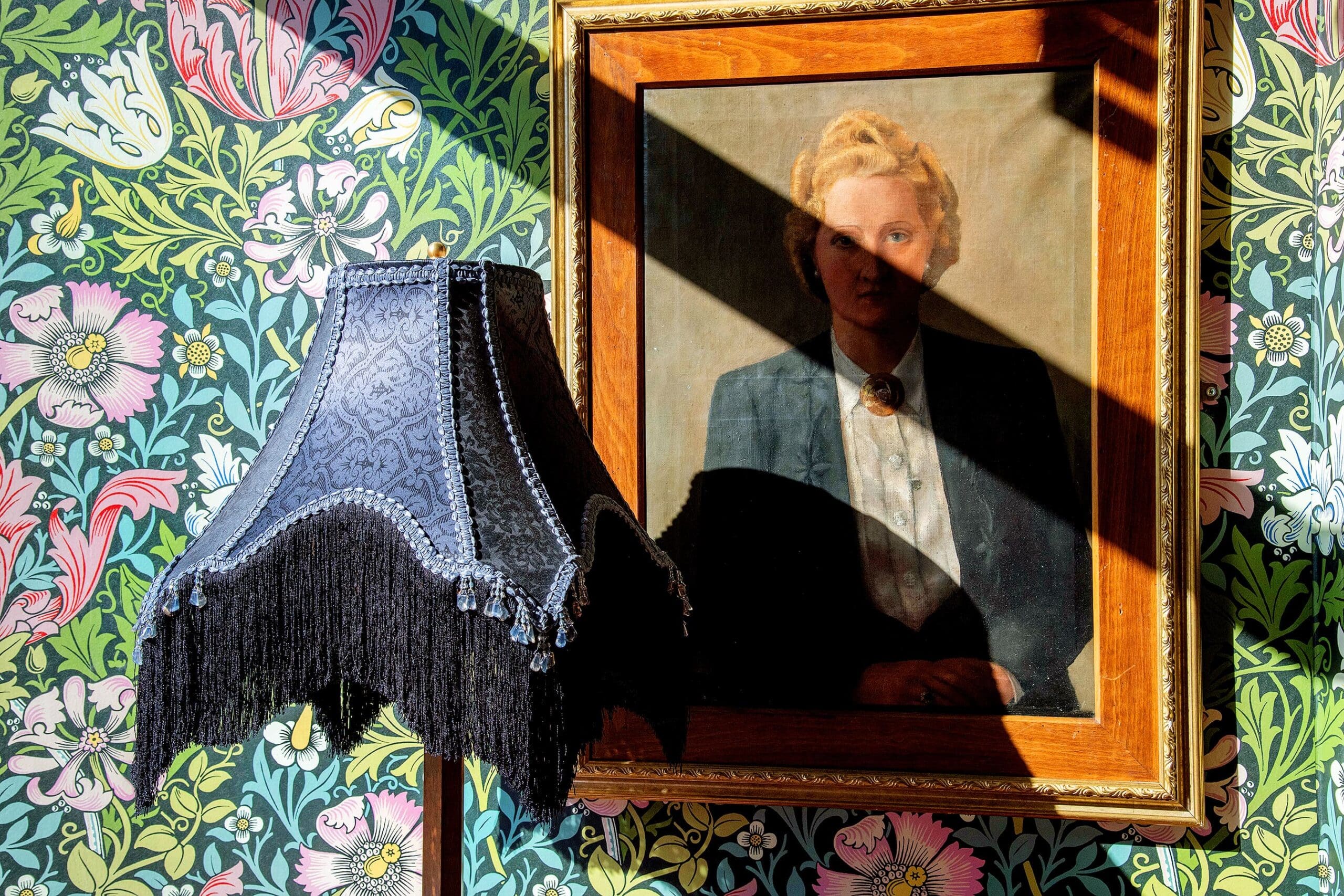A fringed, dark lampshade stands beside a framed portrait of a woman in a suit, both set against vibrant floral wallpaper with subtle ovino motifs. A bright sunlight beam casts a diagonal shadow across the painting.