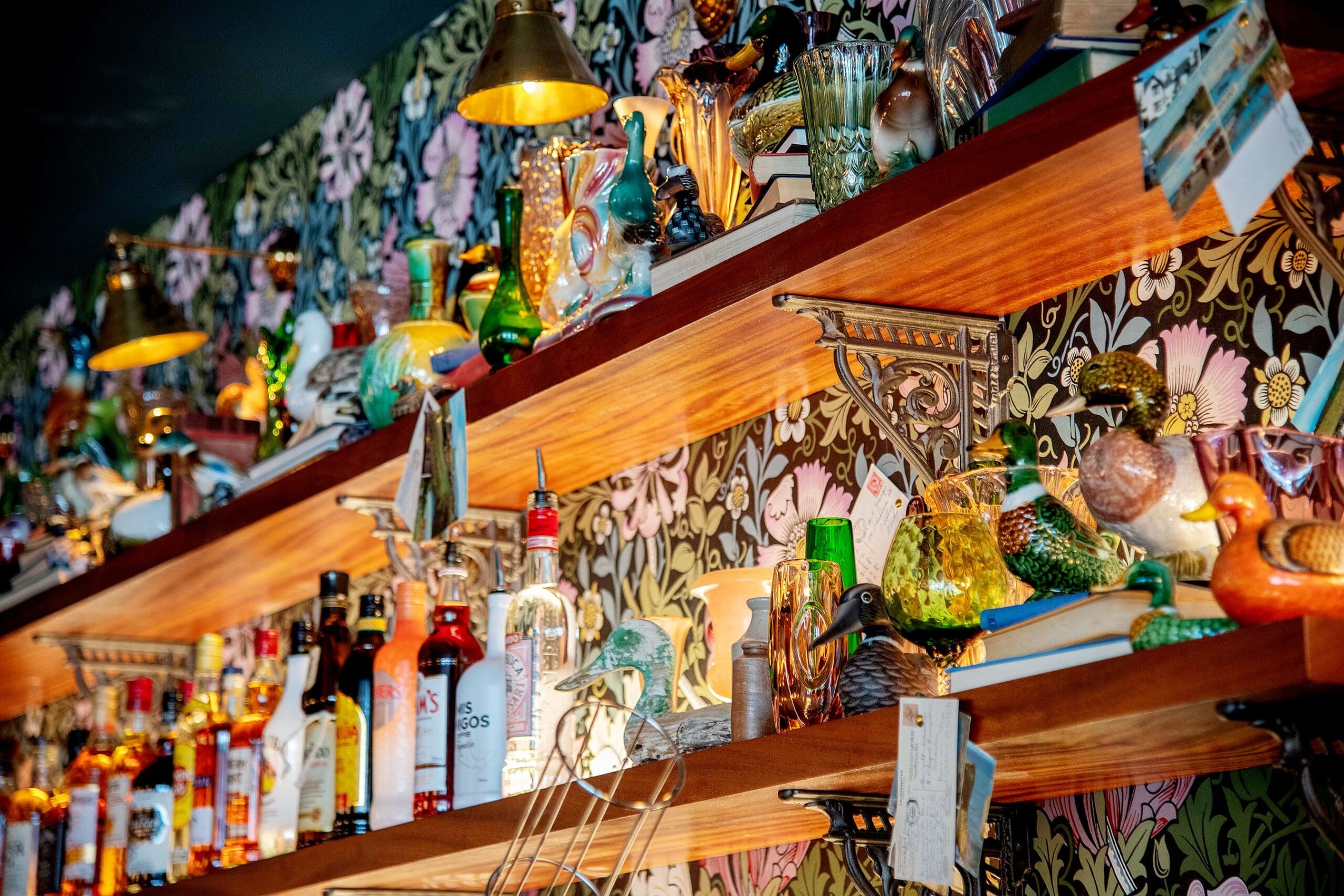 Wooden shelves lined with assorted liquor bottles and decorative ceramic birds, set against floral-patterned wallpaper and illuminated by warm, golden light from nearby lamps—an inviting space perfect for savoring an ovino moment.
