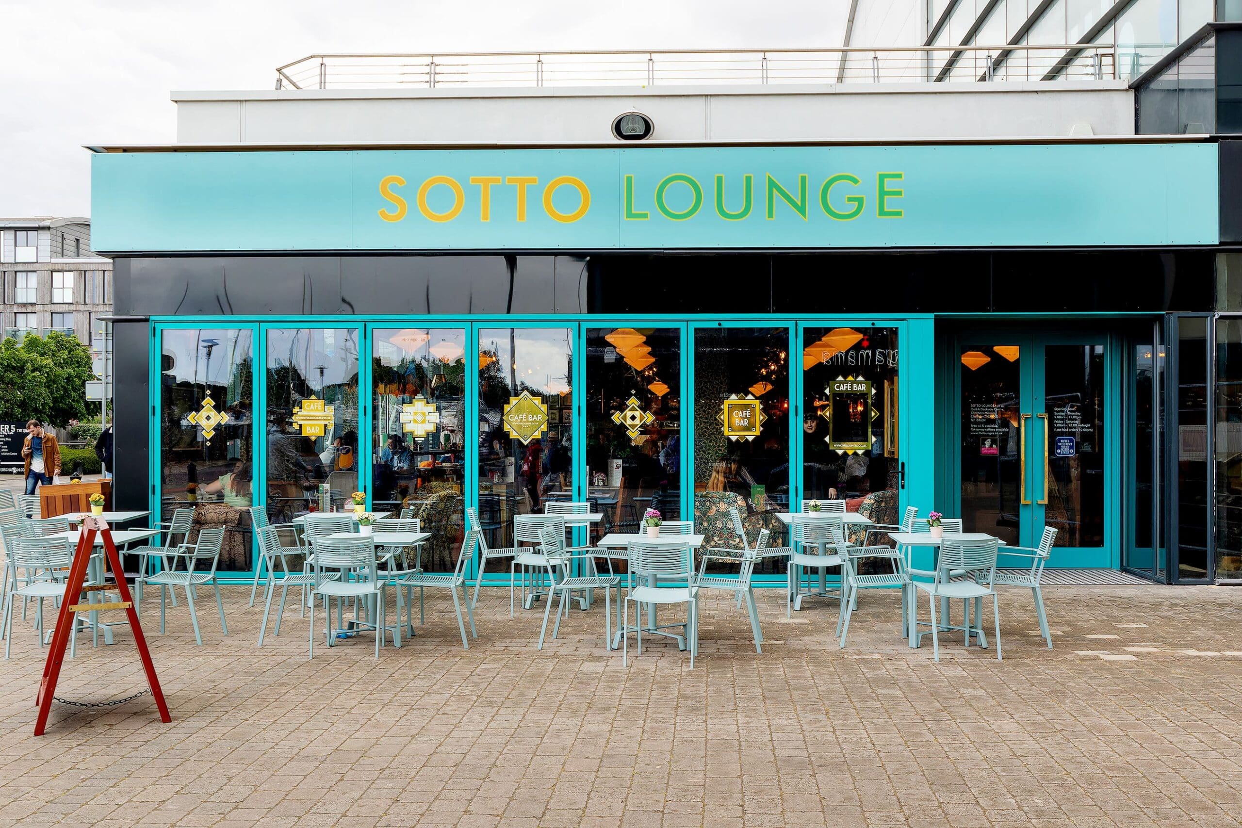 Outdoor seating area with light blue tables and chairs in front of Sotto Lounge café, featuring large glass windows, teal trim, and the inviting Sotto sign above the entrance.