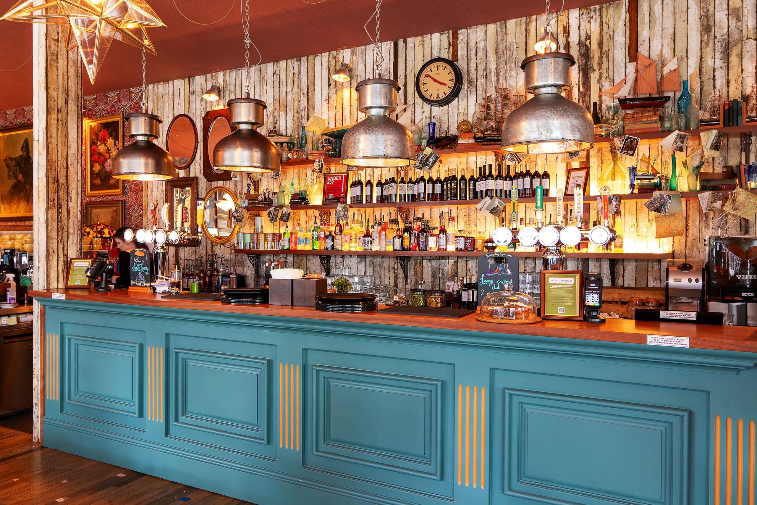 Colorful Sotto bar with a teal counter, hanging industrial lamps, and shelves filled with bottles, glasses, and eclectic decorations against a rustic wooden wall, creating a cozy, vintage atmosphere.