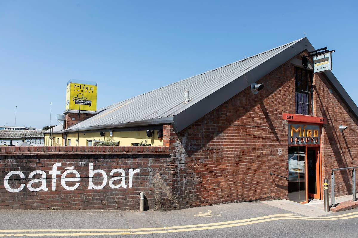 A brick building with a sloped metal roof houses "Miro Lounge," a cozy café bar. Large white "café bar" text and Miro signage stand out on the exterior, making Miro Lounge easy to spot from the street.