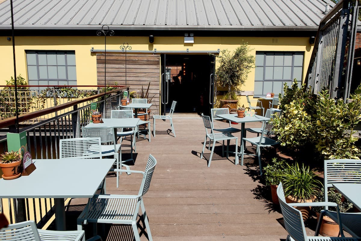 Outdoor patio with light blue tables and chairs, Miro-inspired potted plants along the edges, and a wooden walkway leading to an open door of a yellow building with large windows.