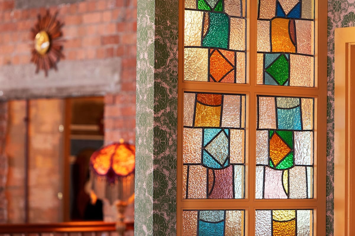 A close-up of a decorative stained glass window with colorful geometric patterns inspired by Miro. In the background, a room features exposed brick walls, a vintage-style lamp, and a sunburst wall clock.