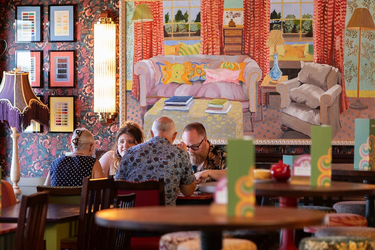 Four people sit around a table in a cozy, colorfully decorated restaurant. Behind them is a large, vibrant Miro-inspired painting of a living room with a sofa, armchair, and window view. The atmosphere appears warm and relaxed.