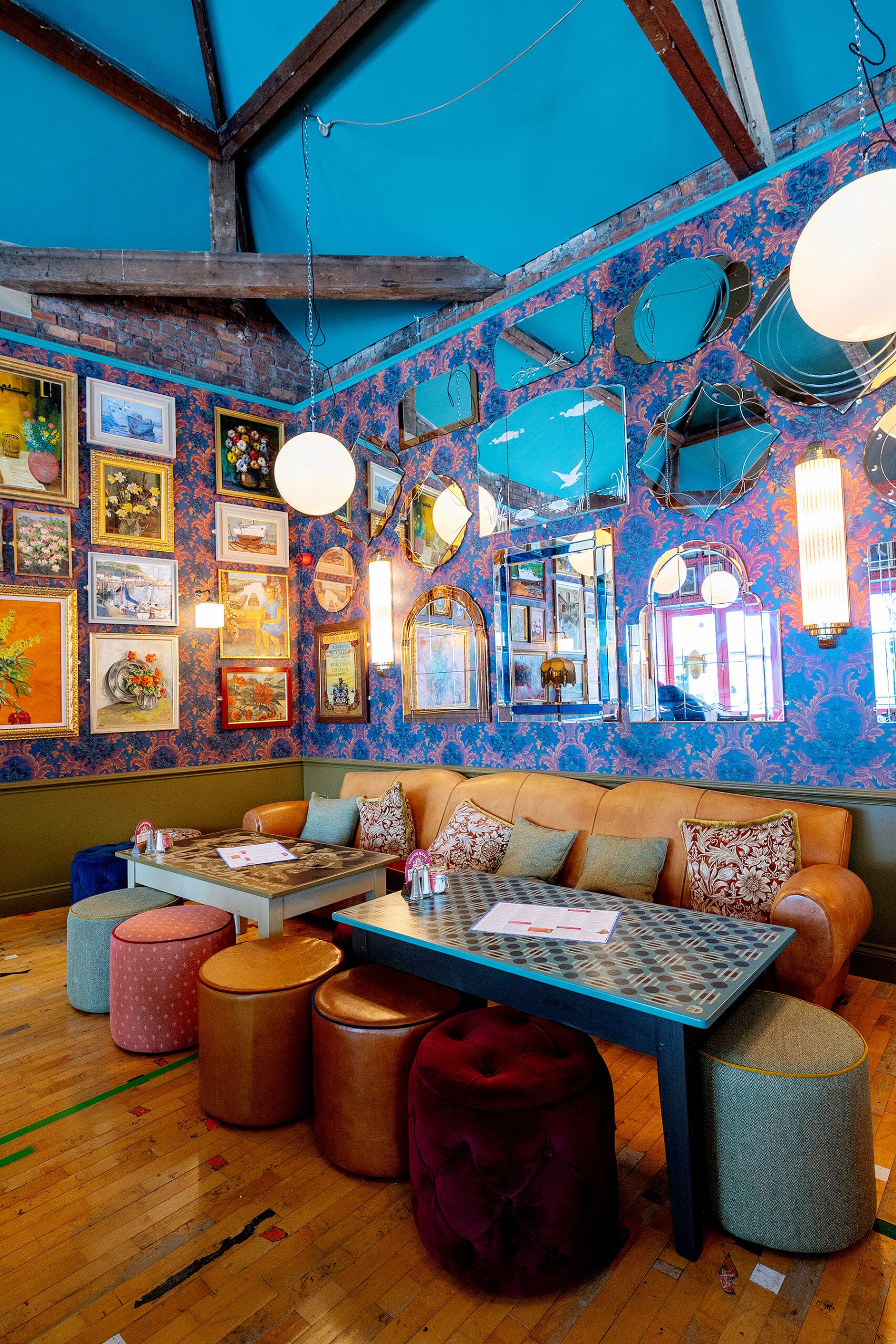 A vibrant Piloto lounge with patterned wallpaper, eclectic framed art, round mirrors, and globe pendant lights. Cozy seating includes a tan sofa, colorful poufs, and a blue patterned table on a wooden floor.