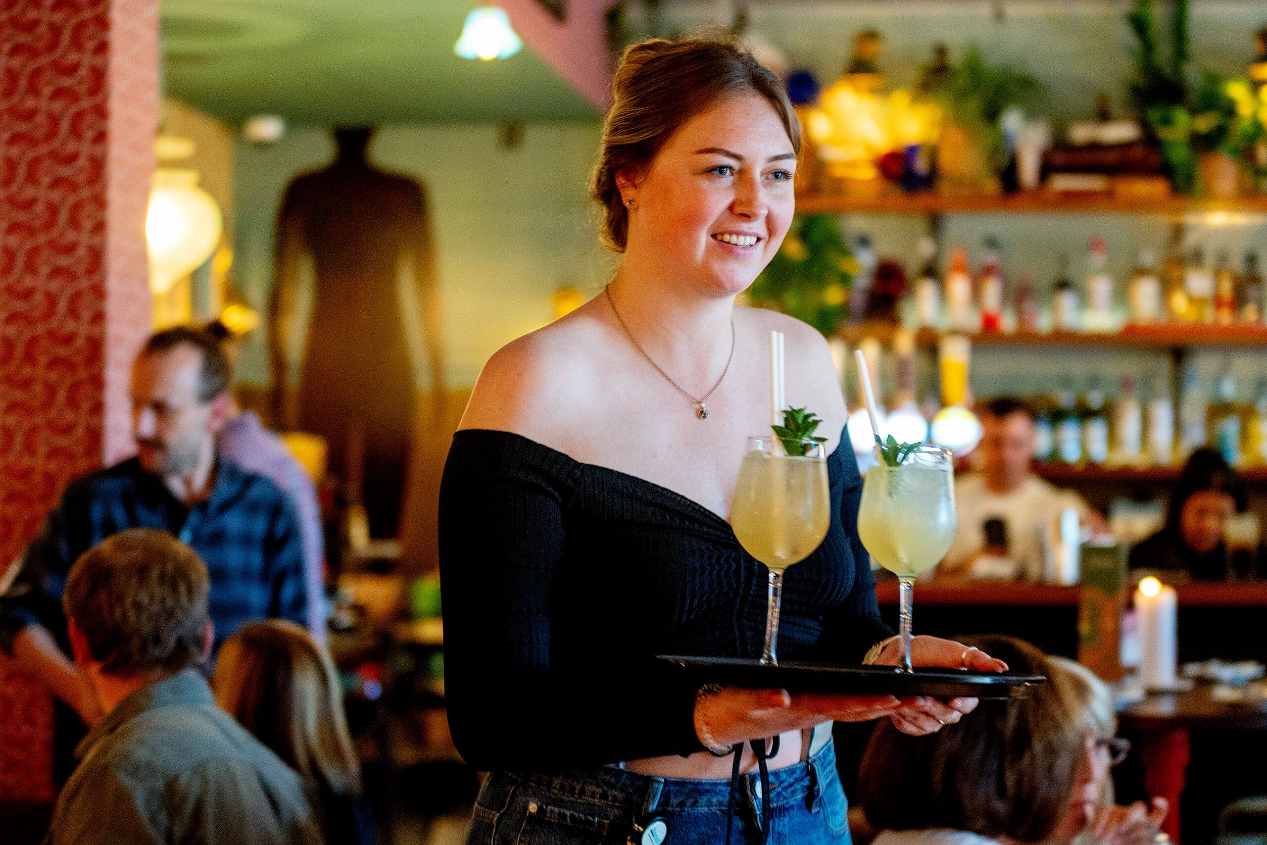 A smiling Piloto waitress in an off-the-shoulder black top carries a tray with two cocktails in a lively, warmly lit restaurant filled with people and shelves stocked with bottles in the background.