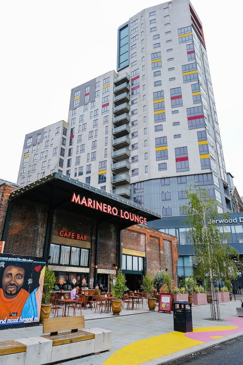 Outdoor view of Marinero Lounge café bar with people dining on the terrace, a colorful mural on the left, and a tall modern apartment building in the background. Trees and planters decorate the scene.