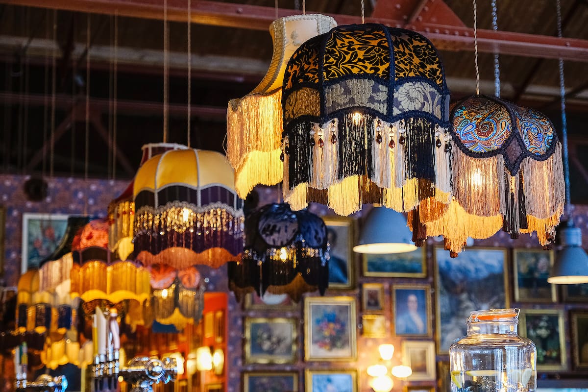 A row of colorful, ornate vintage lampshades with tassels hang from the ceiling in a cozy, eclectic room decorated with framed pictures and marinero-inspired accents under warm lighting.