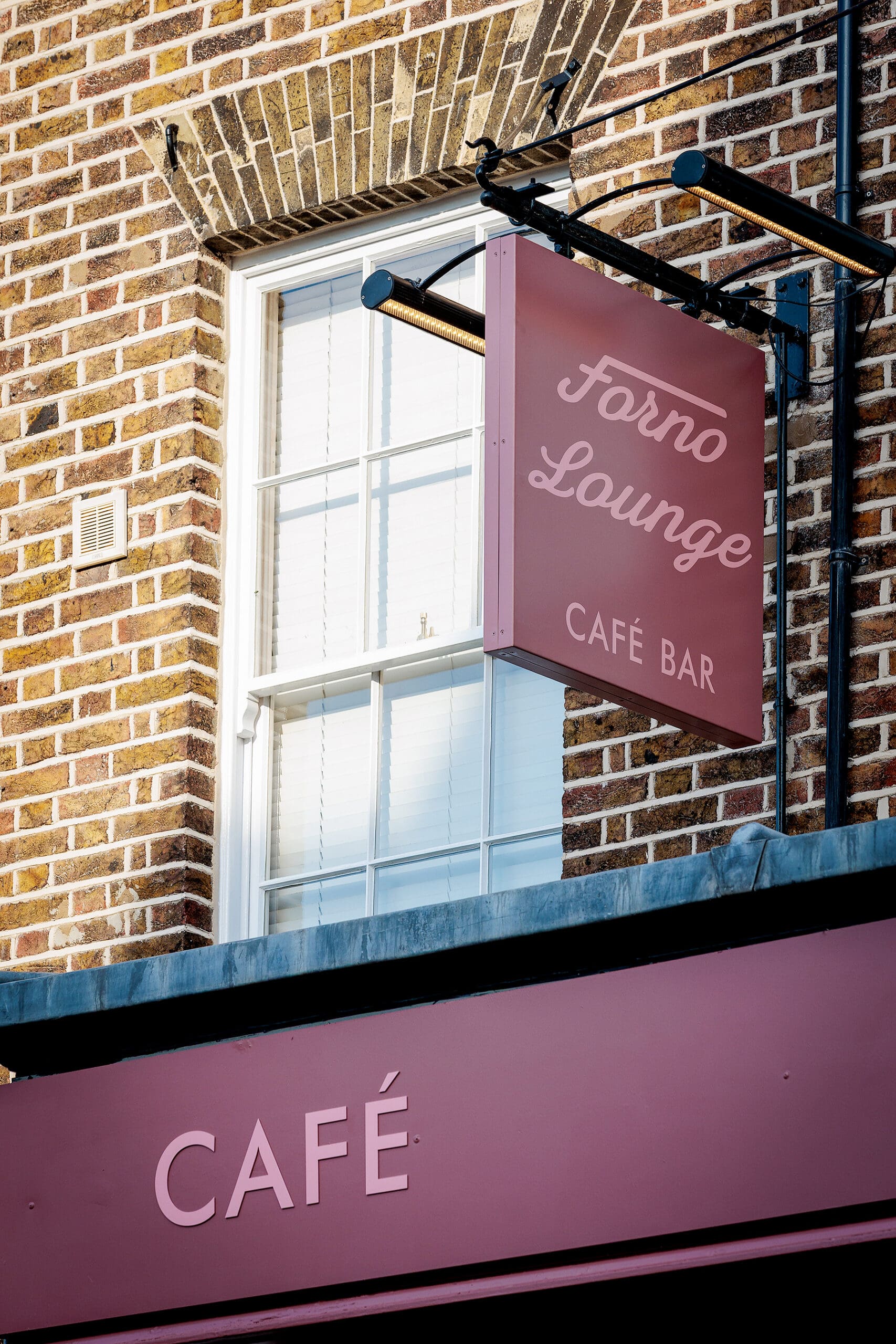 A pink hanging sign reading "Forno Lounge Café Bar" adds charm to the brick building, with another "CAFÉ" sign displayed below, reflecting the inviting forno atmosphere inside.