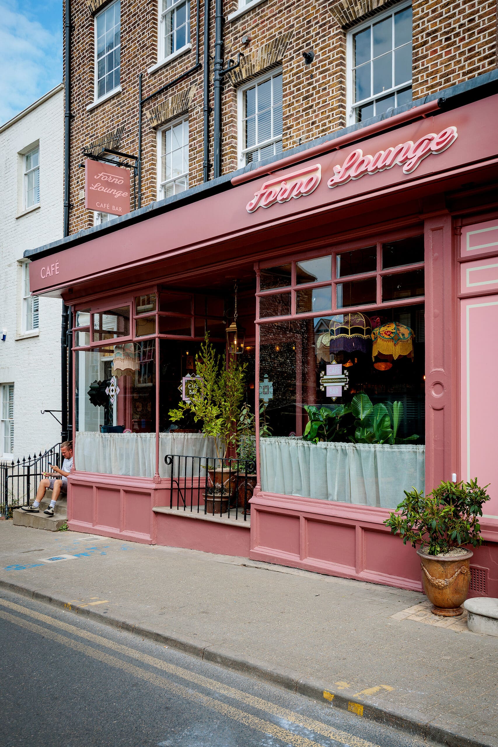 A pink cafe called "Town Lounge" with large windows, potted plants, and neon signage on a brick building featuring a cozy forno inside. A person sits outside on the left, and the street runs in front of the cafe.