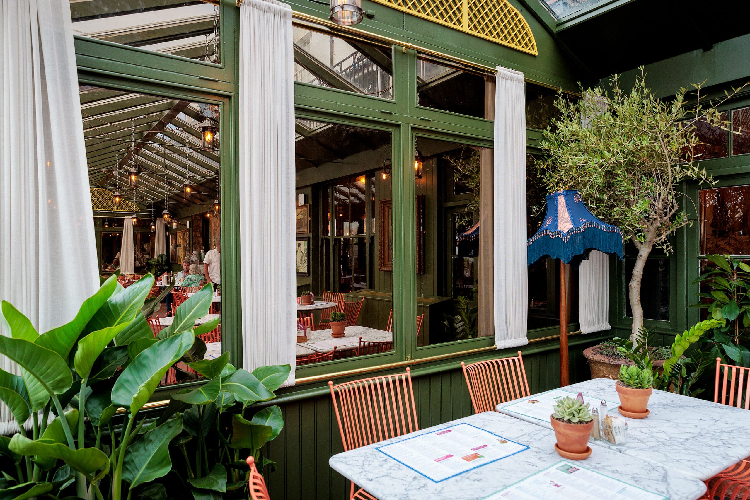 A bright, glass-enclosed restaurant with green-framed windows, marble tables, orange chairs, a forno oven, potted plants, and a blue lamp creates a cozy, inviting atmosphere awash in natural light.