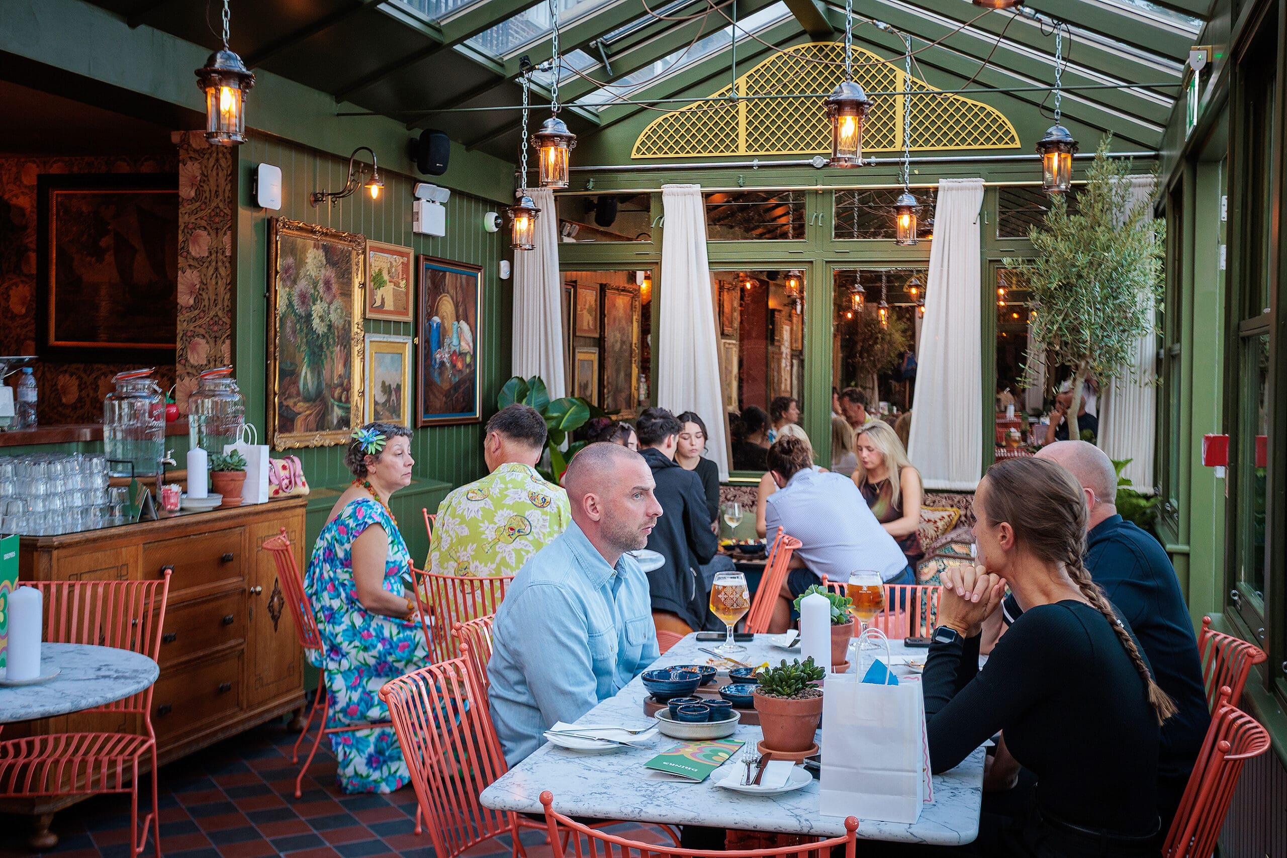 People sit at marble tables in a cozy, plant-filled restaurant with glass ceilings and walls, enjoying forno-baked dishes and conversation. Colorful paintings and warm lights decorate the intimate space.