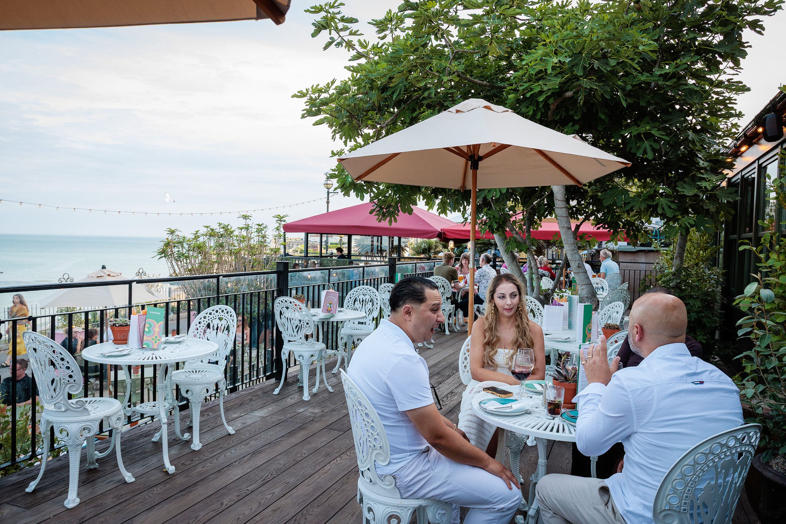 Three people sit at a white metal patio table under an umbrella, enjoying drinks and fresh bites from the forno at a seaside restaurant. Other tables, lush greenery, and the ocean are visible in the background.