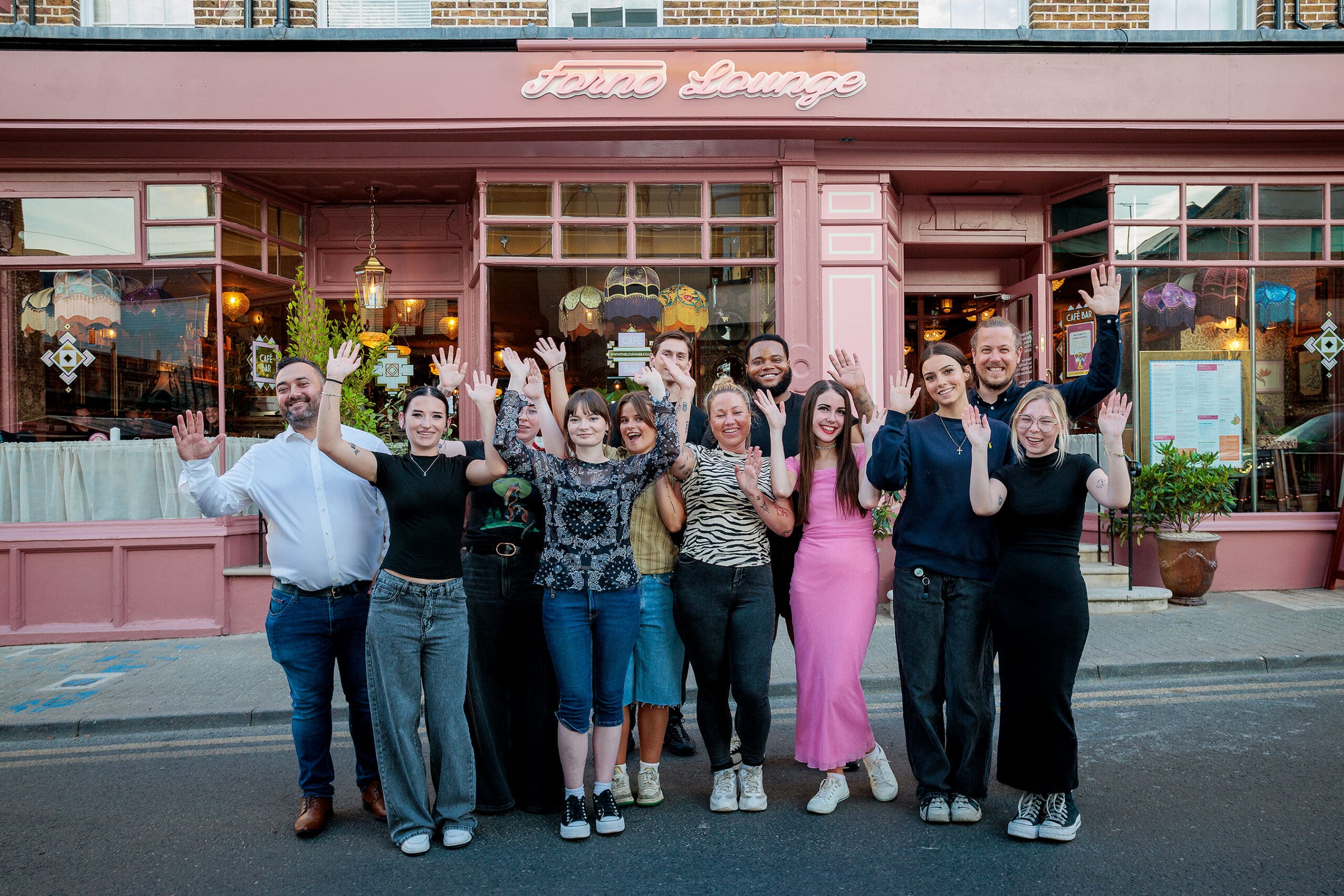 A group of ten people stand outside a pink restaurant called Forno, smiling and waving at the camera. The restaurant features large windows, hanging lights, and decorative details. The group appears cheerful and friendly.