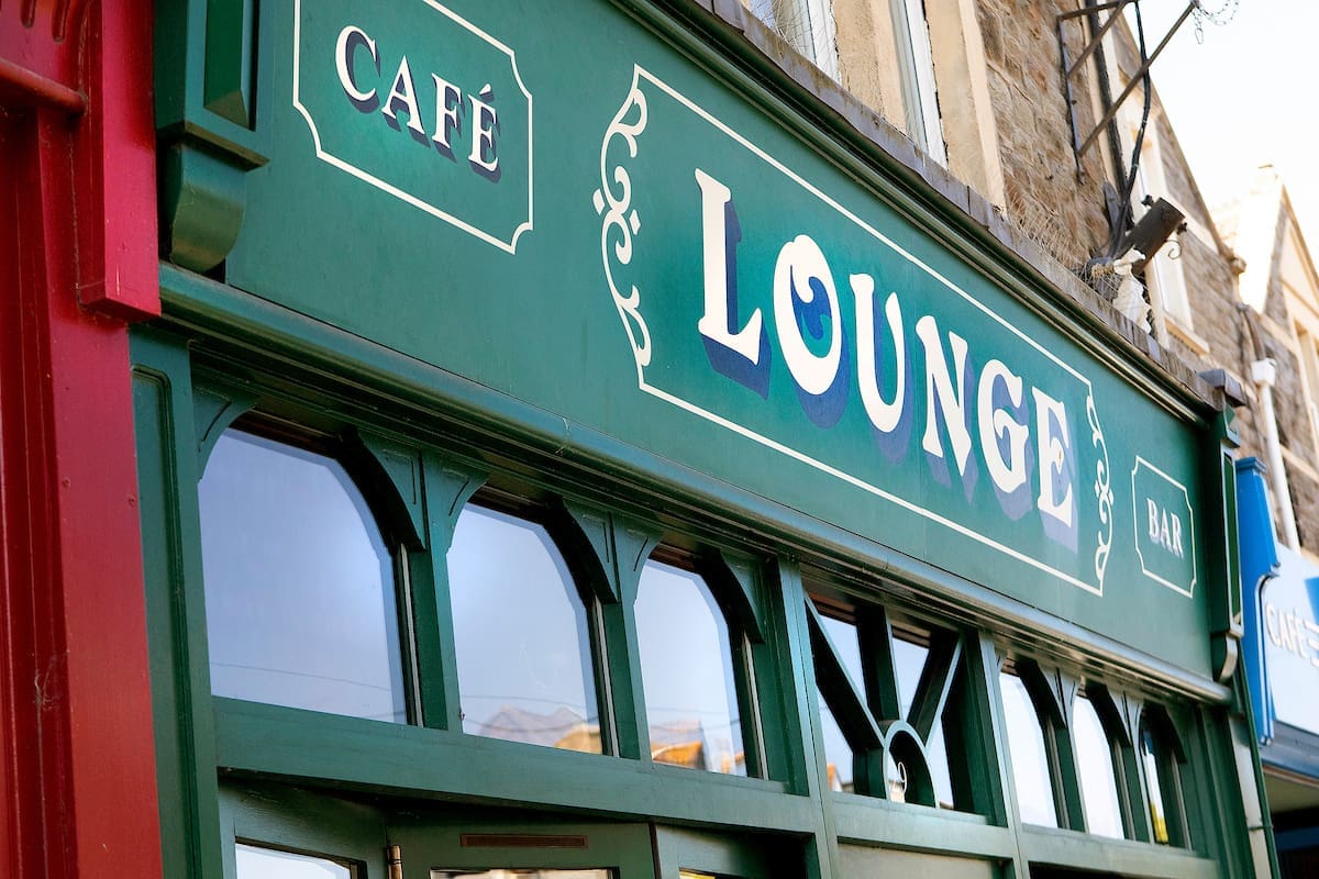 A close-up of the exterior of a green building with signs reading "CAFÉ" and "The Lounge" in white letters. Large windows reflect light, and part of a stone wall is visible above the storefront.