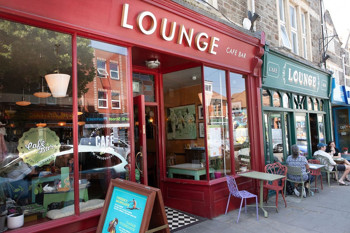 The exterior of The Lounge café bar features a red facade. Outdoor tables host people chatting, while the neighboring café with its green facade contrasts nearby. Reflections and decor are visible through The Lounge’s windows.
