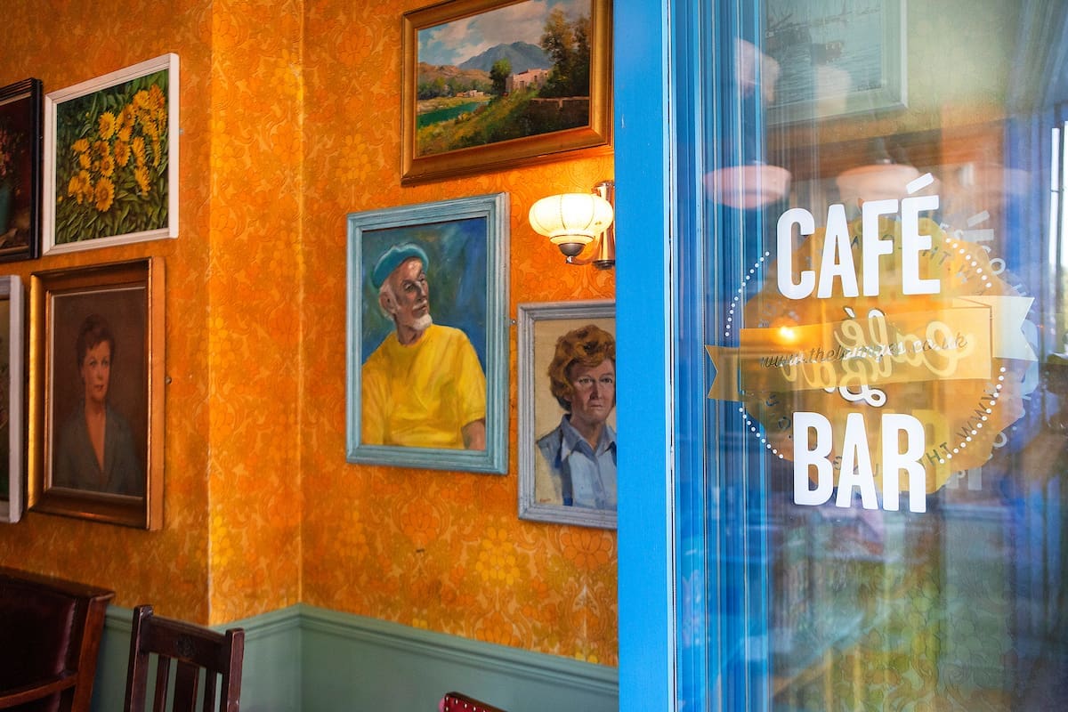 A cozy cafe bar, The Lounge, features bright orange walls adorned with framed portraits and scenic paintings, seen through a glass door labeled "CAFE BAR" in white and gold lettering.