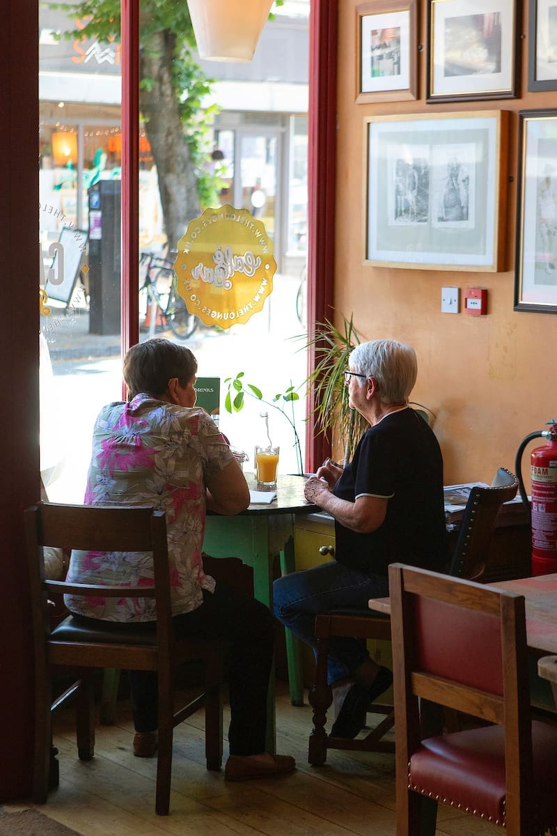 Two older women sit at a small table in The Lounge, a cozy café by a window. They talk and enjoy drinks as sunlight streams through the glass, illuminating plants, artwork, and the lively street outside.