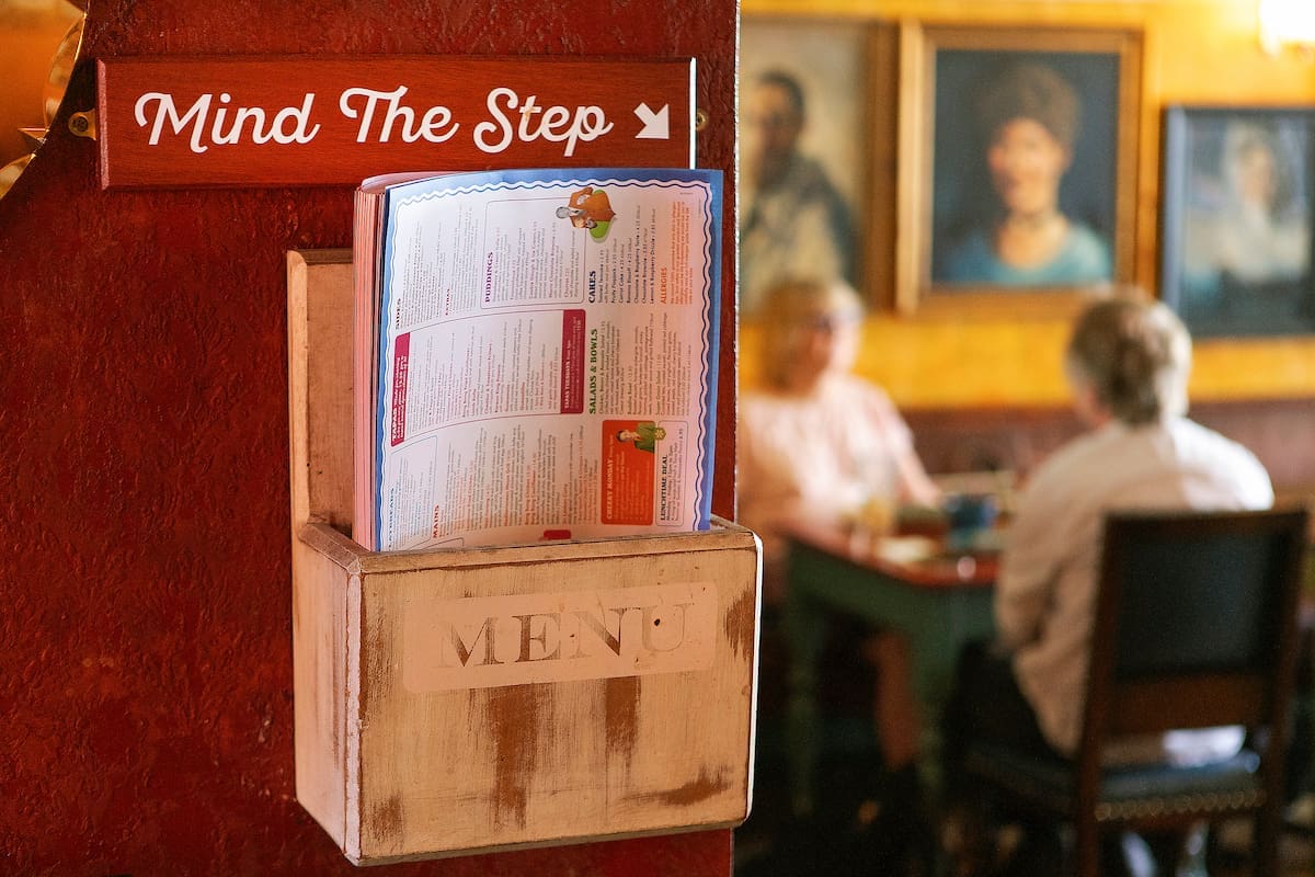 A wooden menu holder with menus and a sign that reads "Mind The Step" on a red wall at The Lounge; two people sit at a table in the background with framed portraits on a yellow wall.
