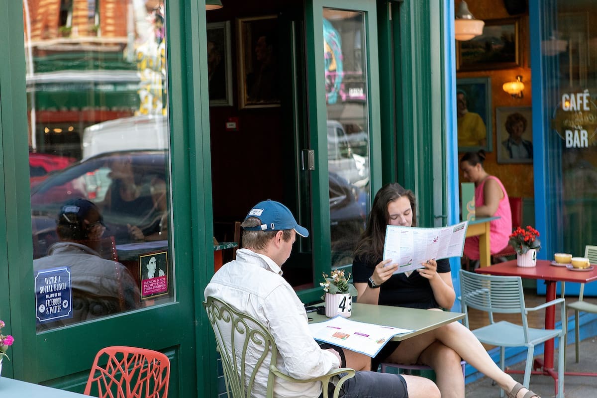 Two people sit at an outdoor café table reading menus at The Lounge. A woman in a pink top stands near the entrance, and the café’s colorful interior and large windows create a casual, urban vibe.