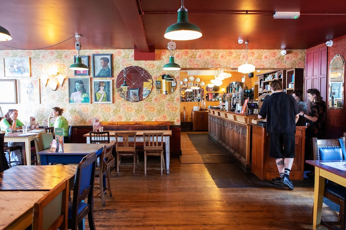 The Lounge features a cozy pub interior with wooden floors, eclectic furniture, floral wallpaper, and various portraits and mirrors on the walls. Two people talk at the bar while others relax at tables near the windows, all bathed in warm lighting.
