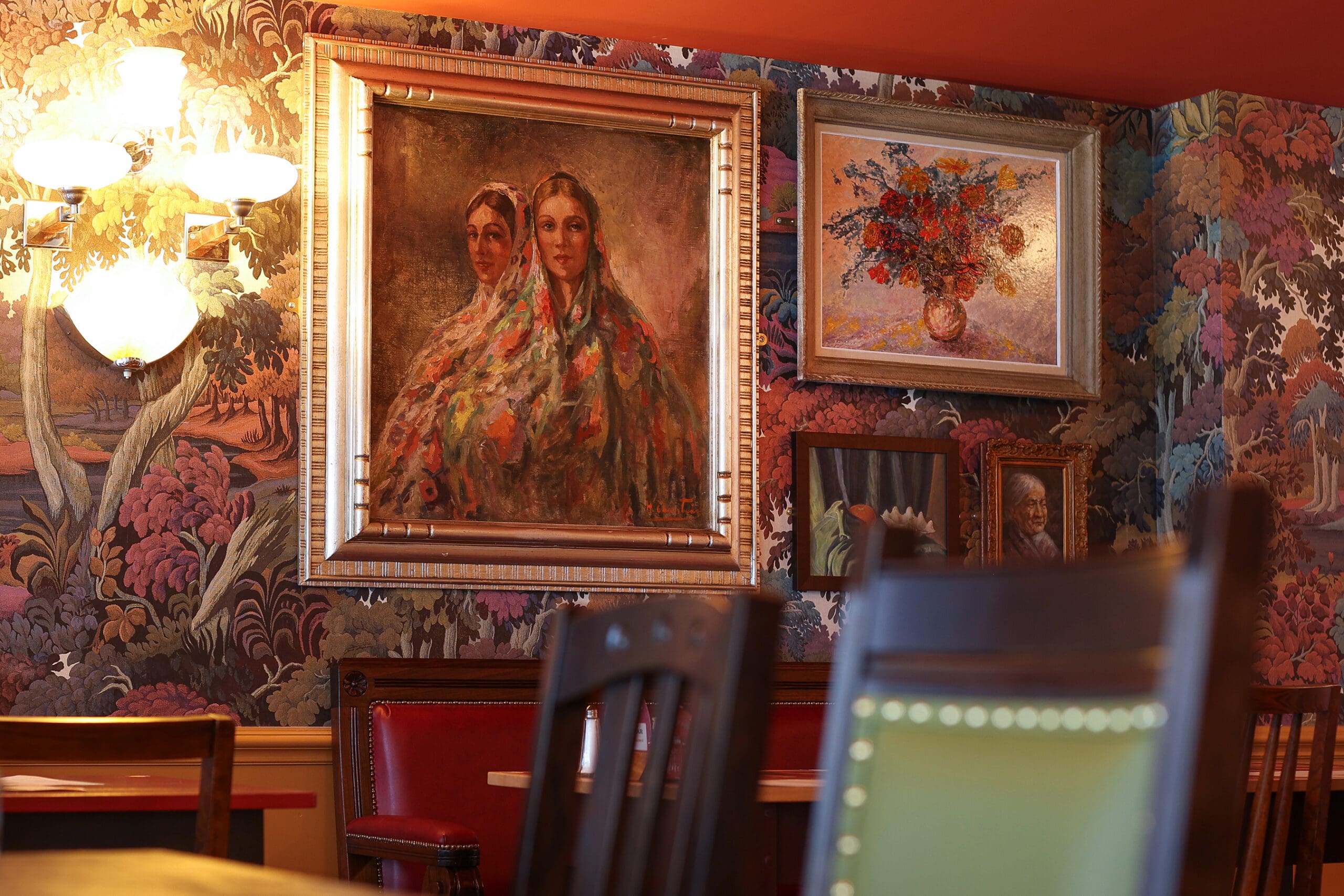 Wall adorned with colorful floral wallpaper, Antico-style framed paintings of women in shawls, a vase of flowers, and portraits; warm lighting with part of a chandelier visible, and wooden chairs and tables in the foreground.