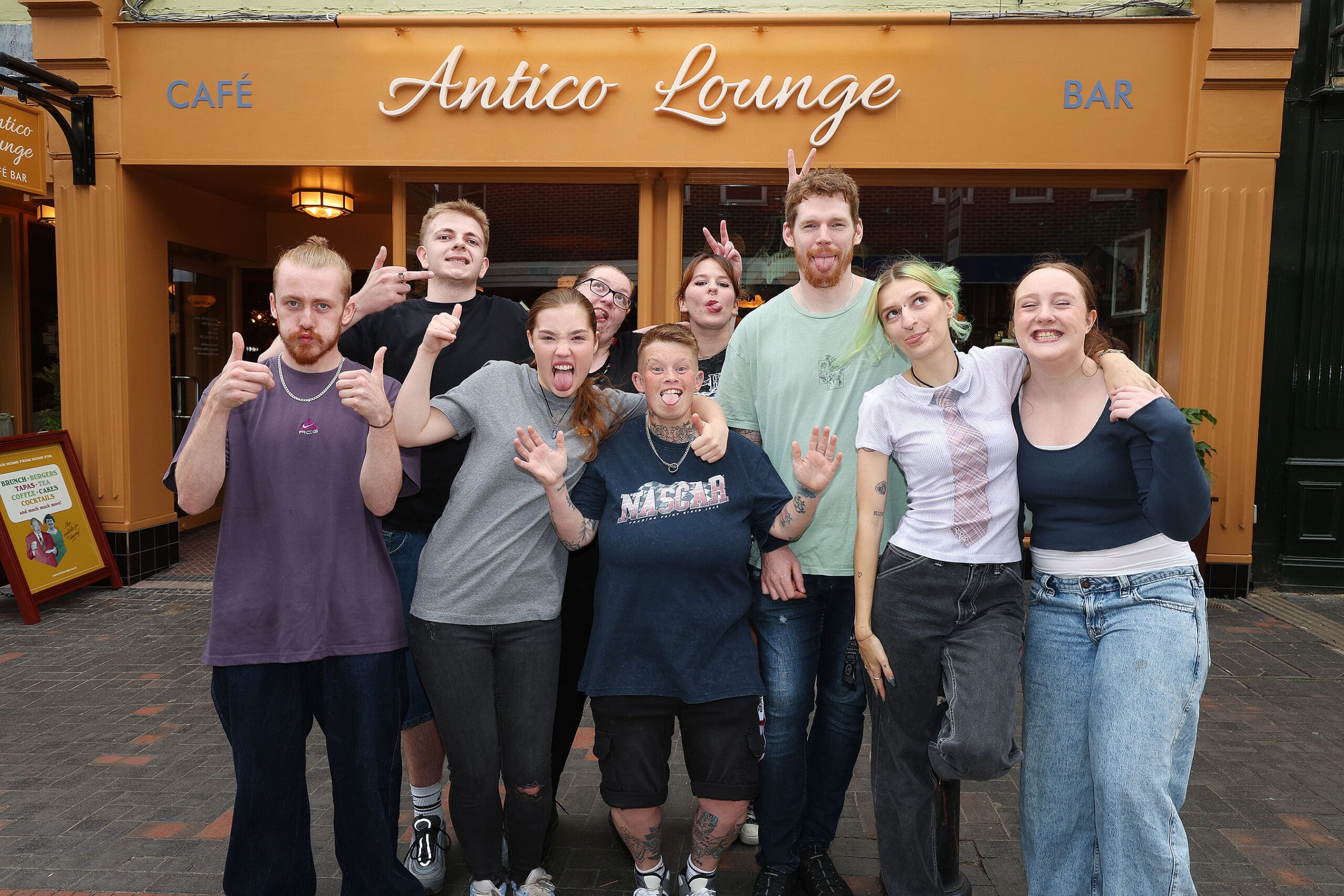 A group of eight smiling people pose in front of Antico, the popular café and bar, standing closely together, some giving thumbs up or peace signs, appearing happy and relaxed.